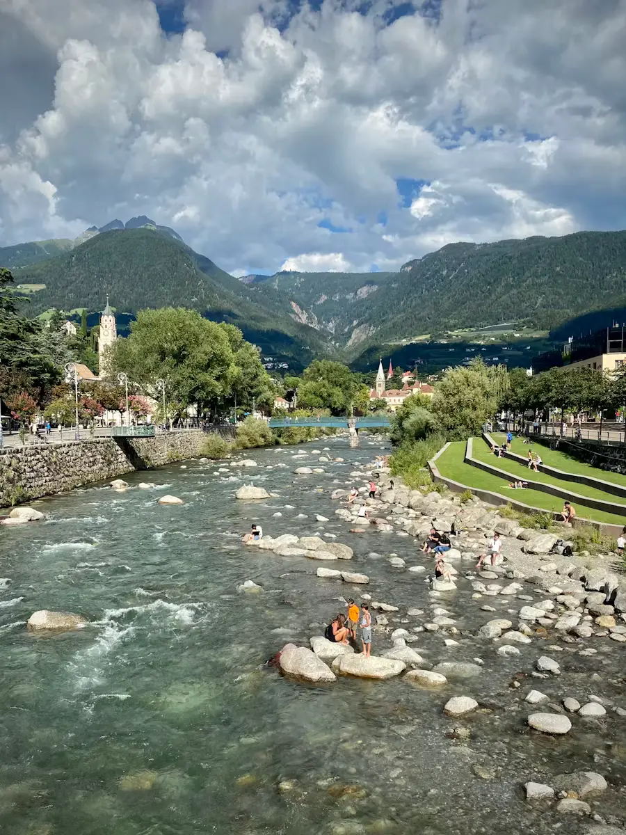 A shallow, rocky river winding through a scenic valley, with people wading near the water's edge and mountains rising in the background.