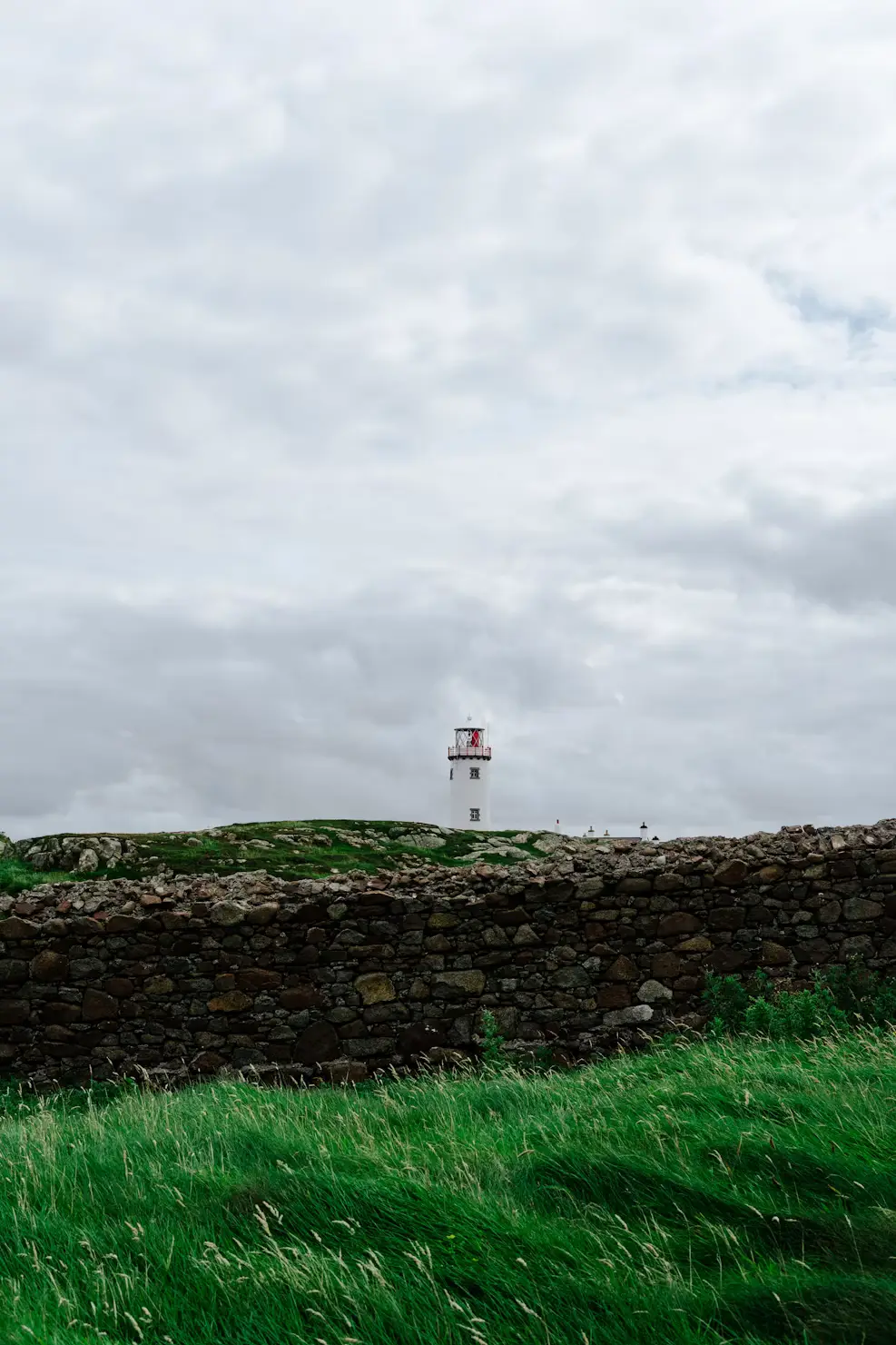 Small distant lighthouse behind a stone wall on a green grassy field, under an overcast sky, Ireland.