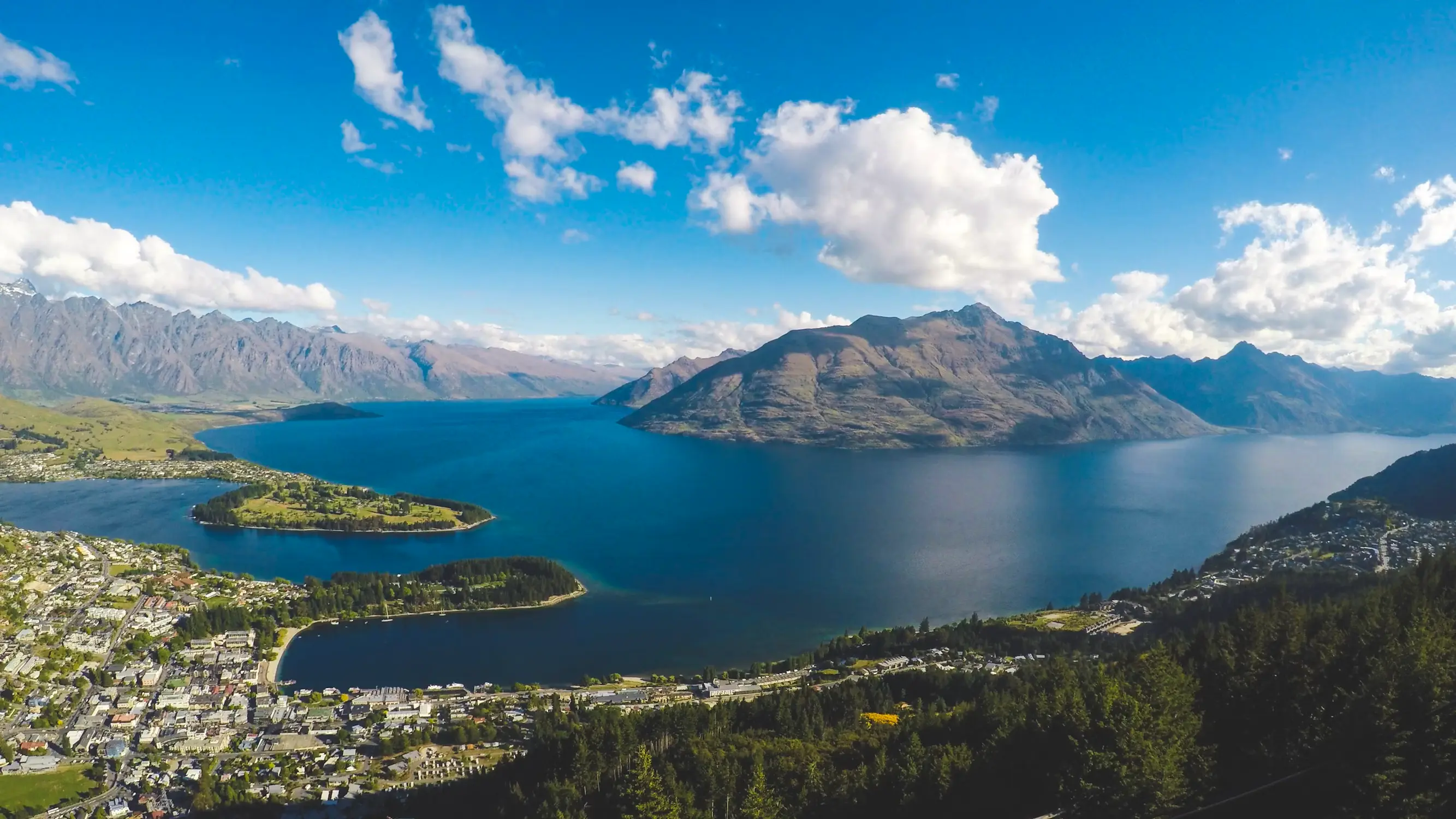 Panoramic view of Lake Wakatipu with forested hills, mountain peaks, and Queenstown beneath a bright blue sky with scattered clouds.