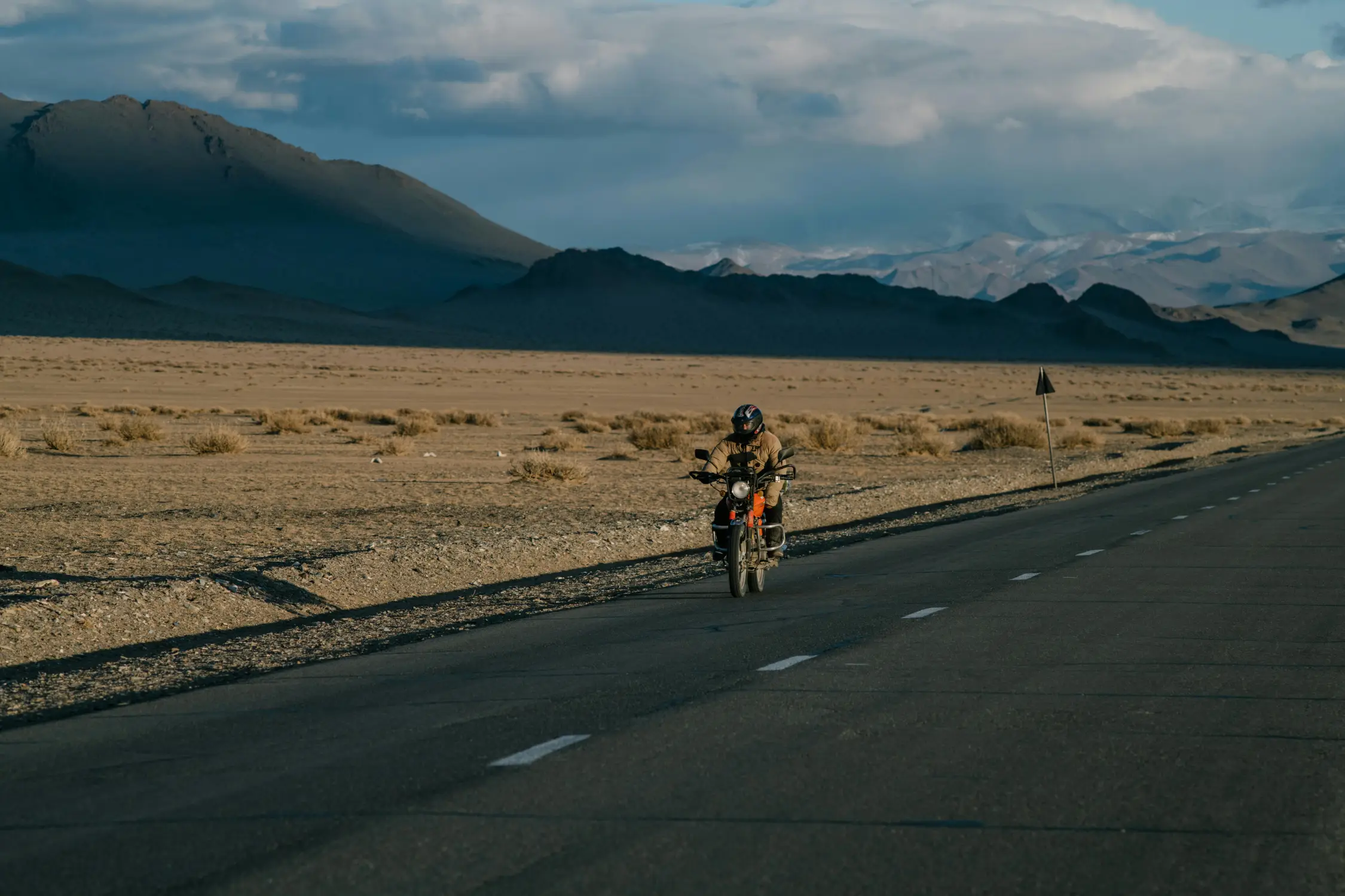 Adventure motorcyclist in full gear riding on a remote desert highway in western Mongolia with dramatic dark mountains and cloudy sky in the background
