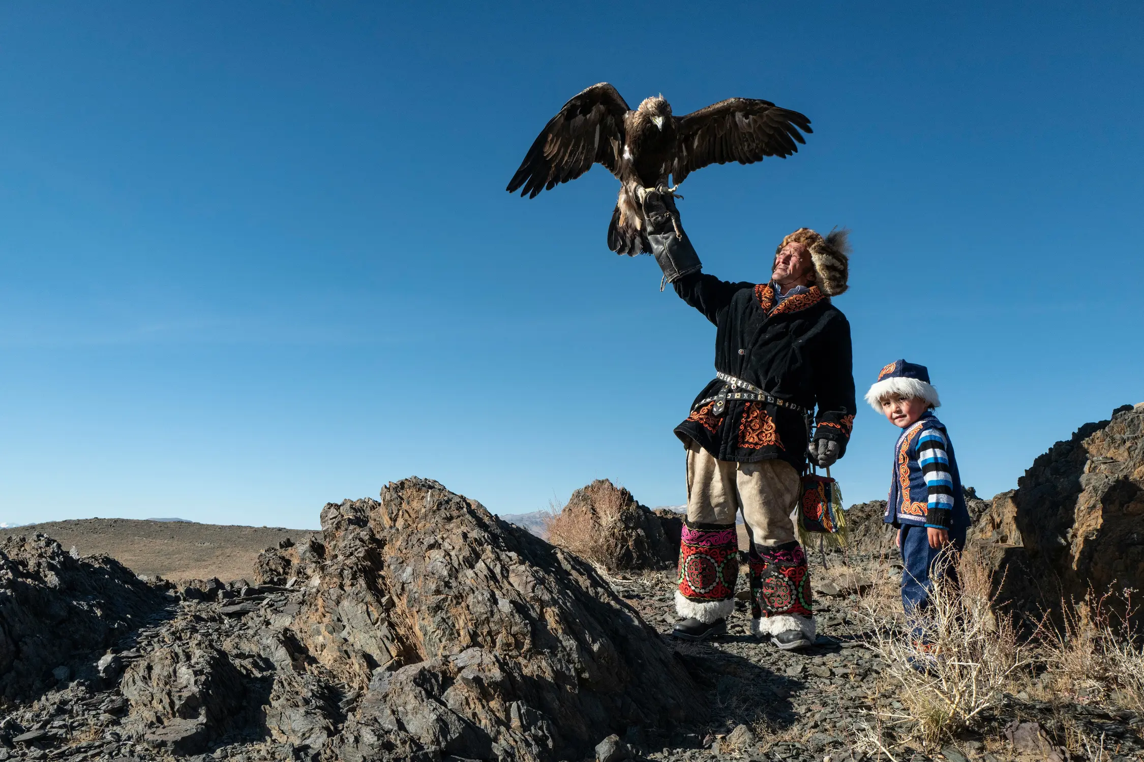 A traditionally dressed Kazakh eagle hunter holding a large golden eagle with outstretched wings on rocky terrain, with a young boy in a white hat standing beside him