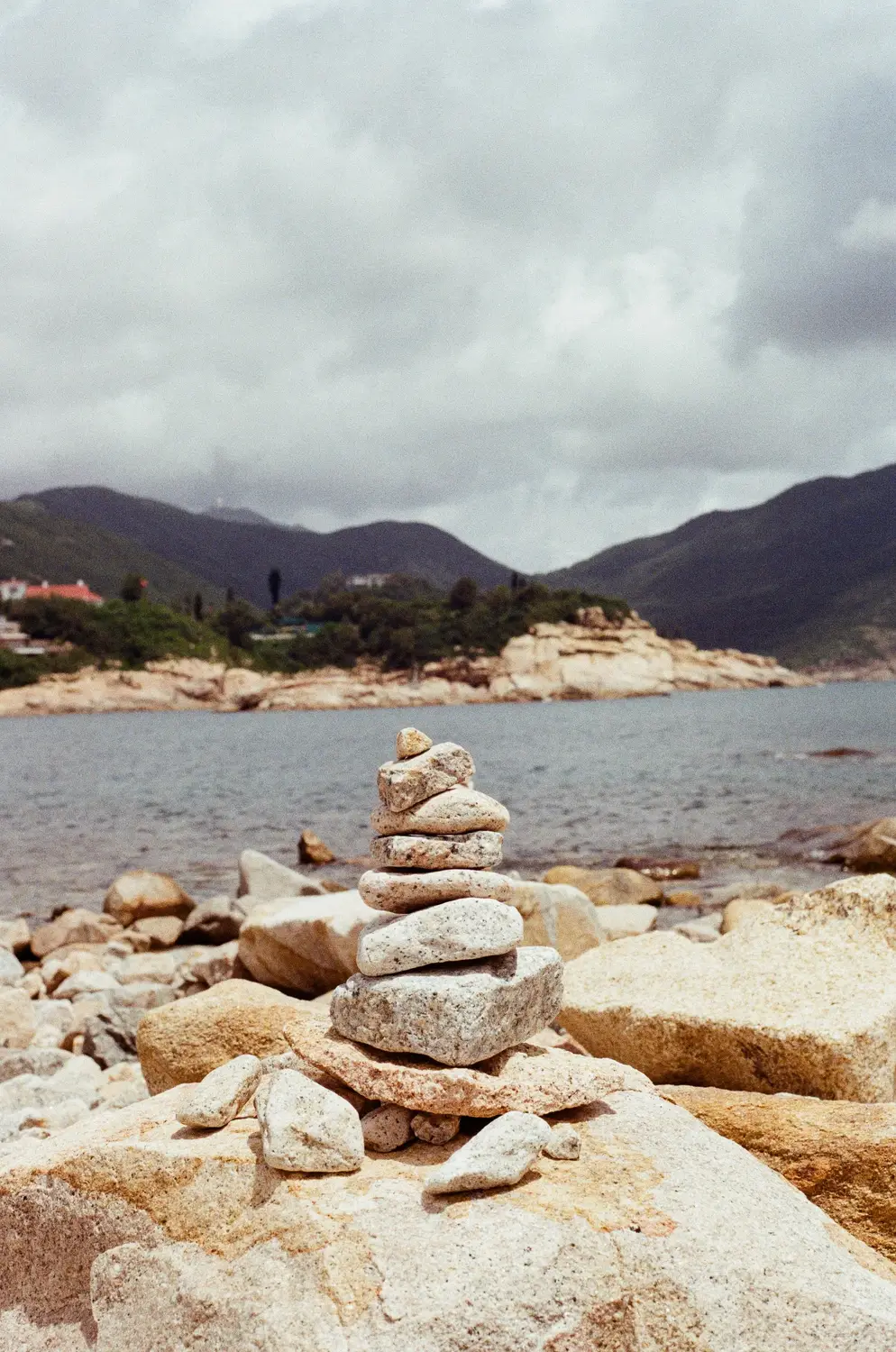 Stacked stone cairns on a rocky beach with mountains and cloudy skies in Hong Kong.
