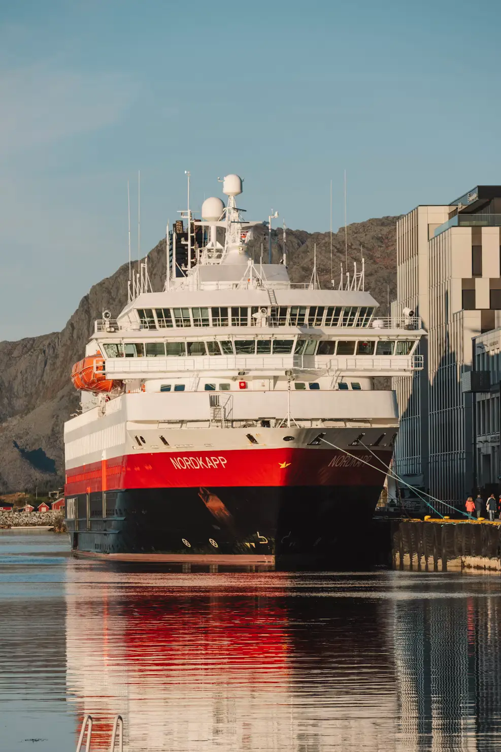 A red and black Hurtigruten cruise ship docked at a calm harbour, its hull reflected in the still water.