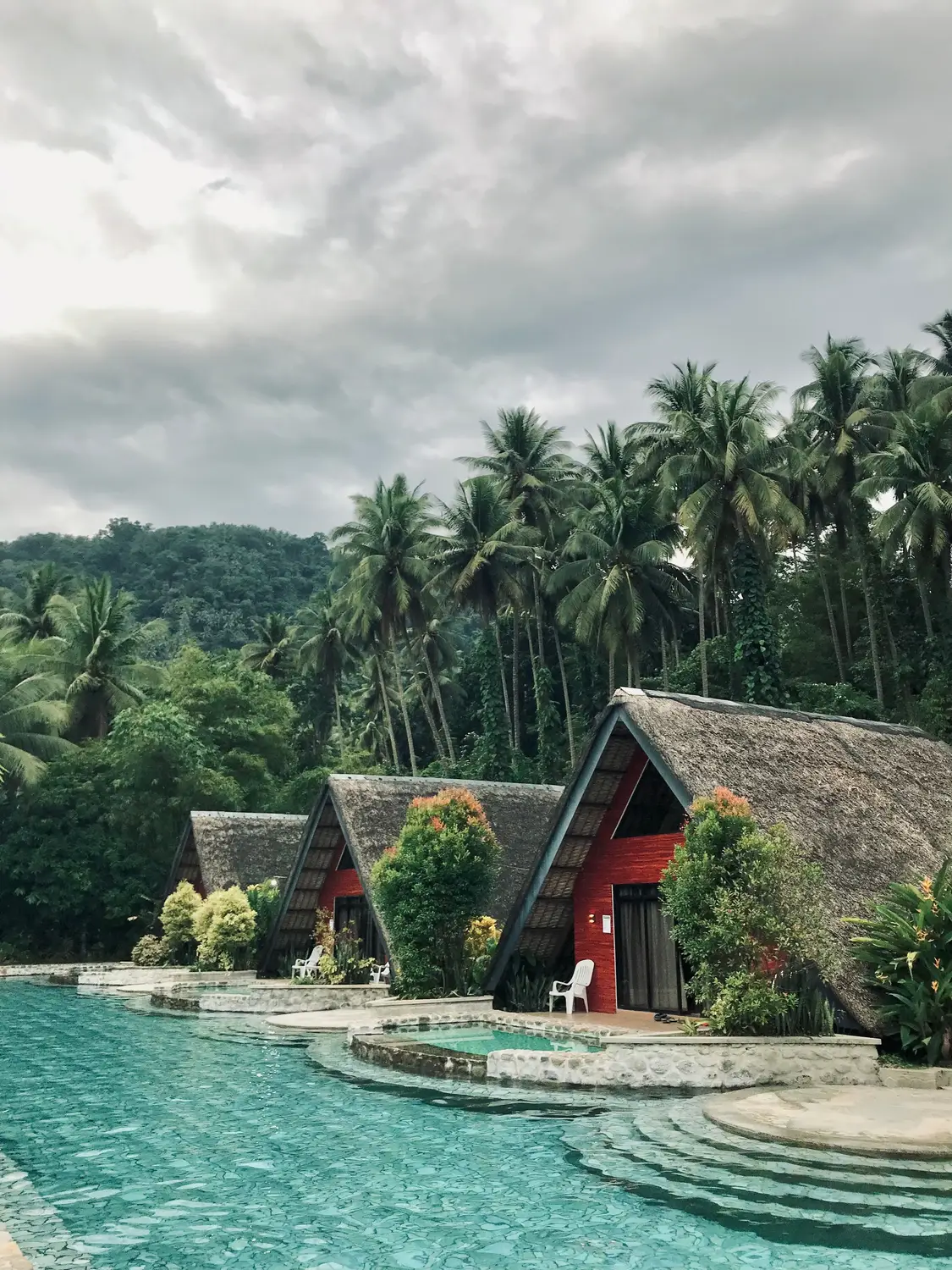 Red A-frame thatched-roof cabins beside a turquoise swimming pool surrounded by palm trees and jungle at a tropical resort in the Philippines.