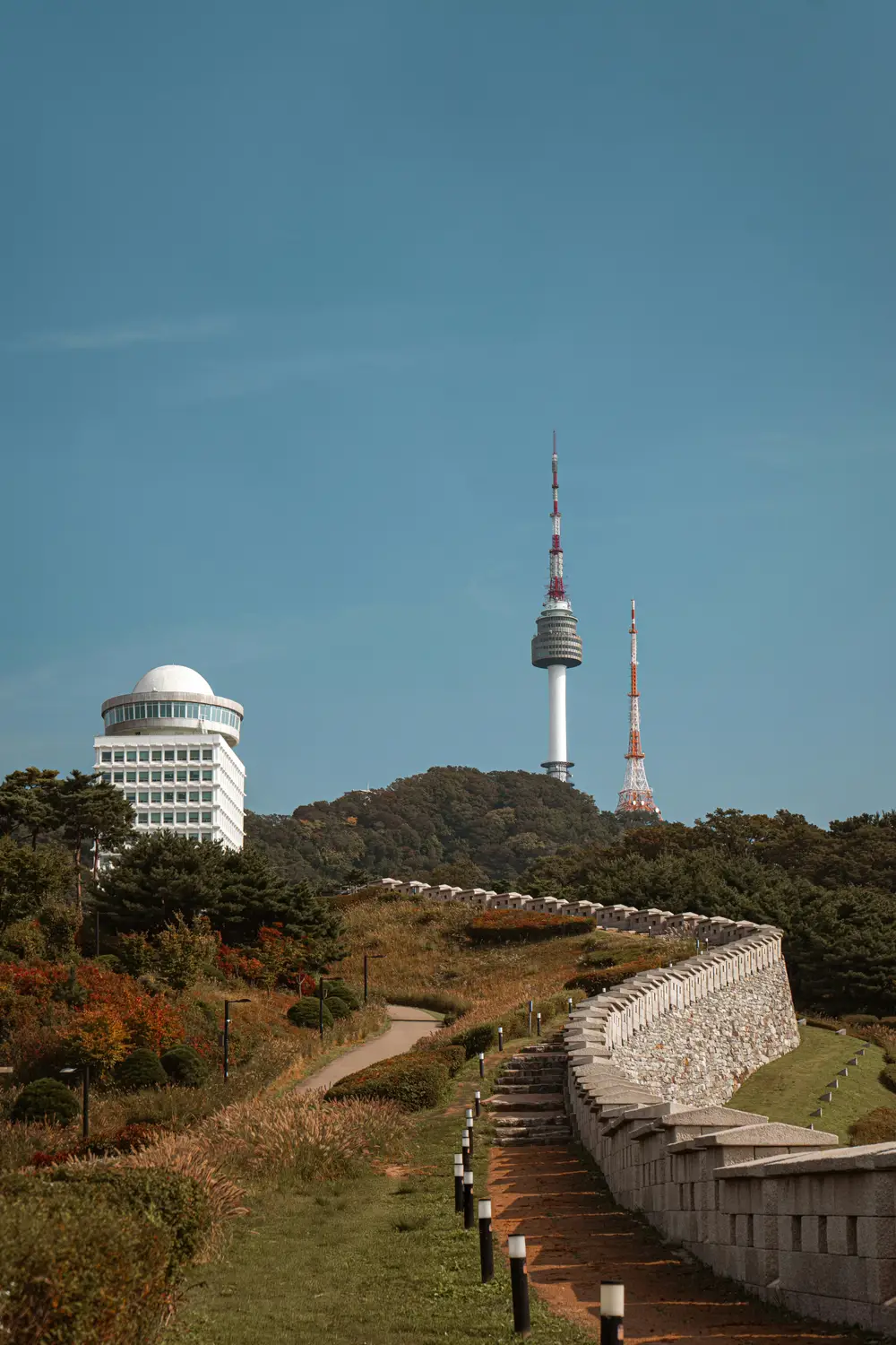 N Seoul Tower rising above the forested slopes of Namsan Mountain, with the old Seoul fortress wall and a stepped pathway in the foreground under a clear blue sky.