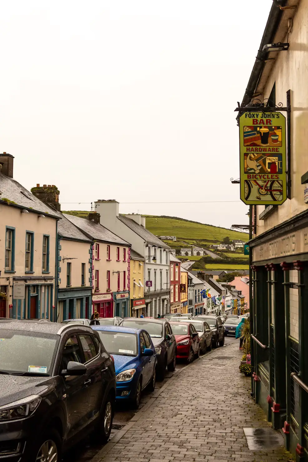Cobblestone street lined with colorful shopfronts and parked cars in a small Irish town at dusk.