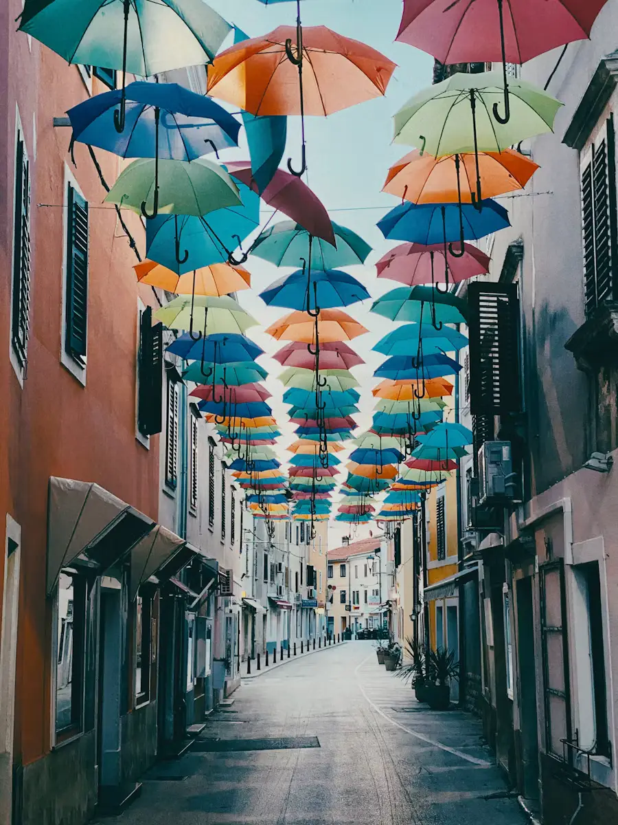 Colorful umbrellas strung overhead above a narrow pedestrian street lined with shops and pastel buildings.