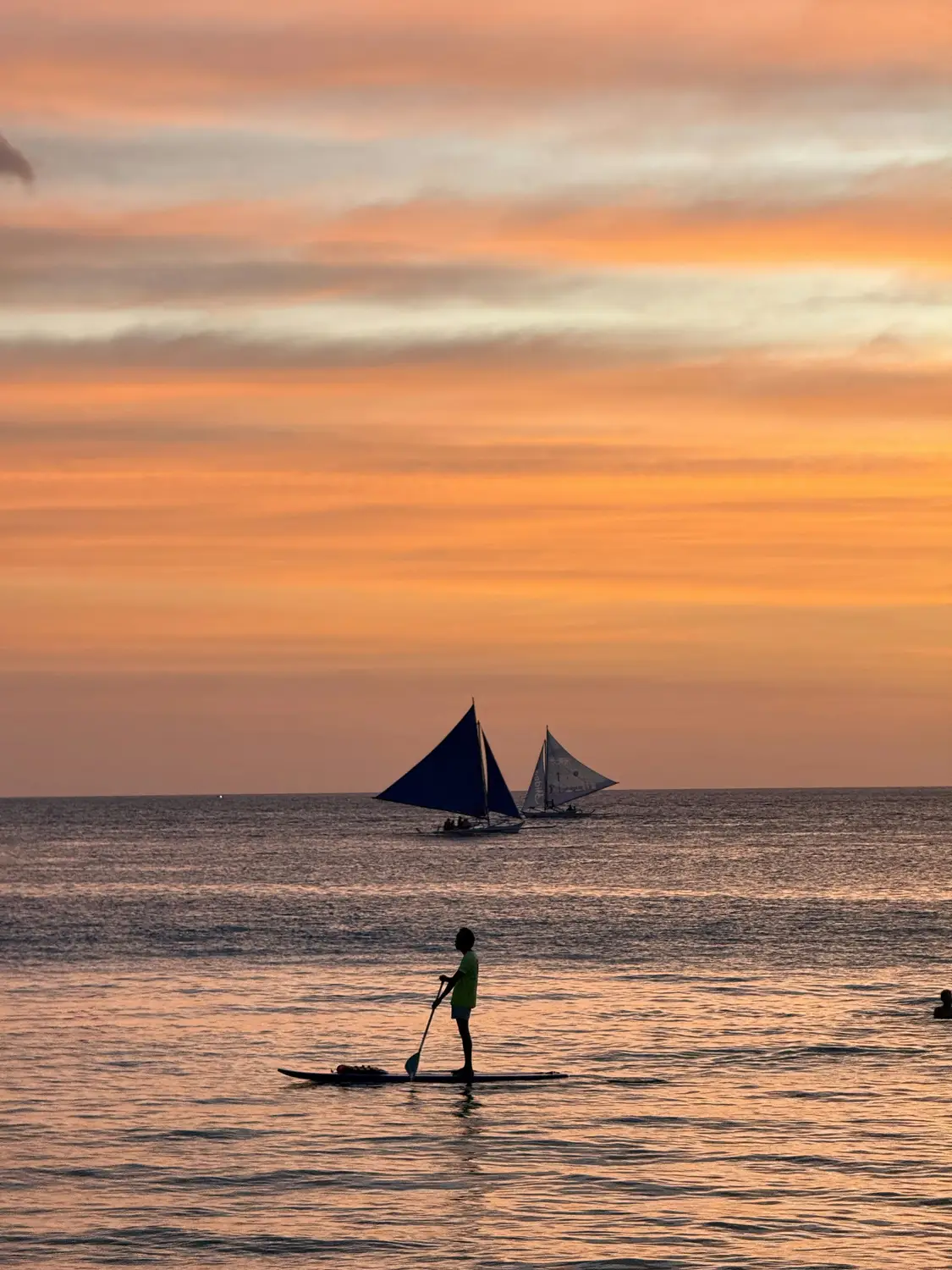 Silhouette of a sailboat and paddleboarder on calm water at sunset in Boracay, Philippines.