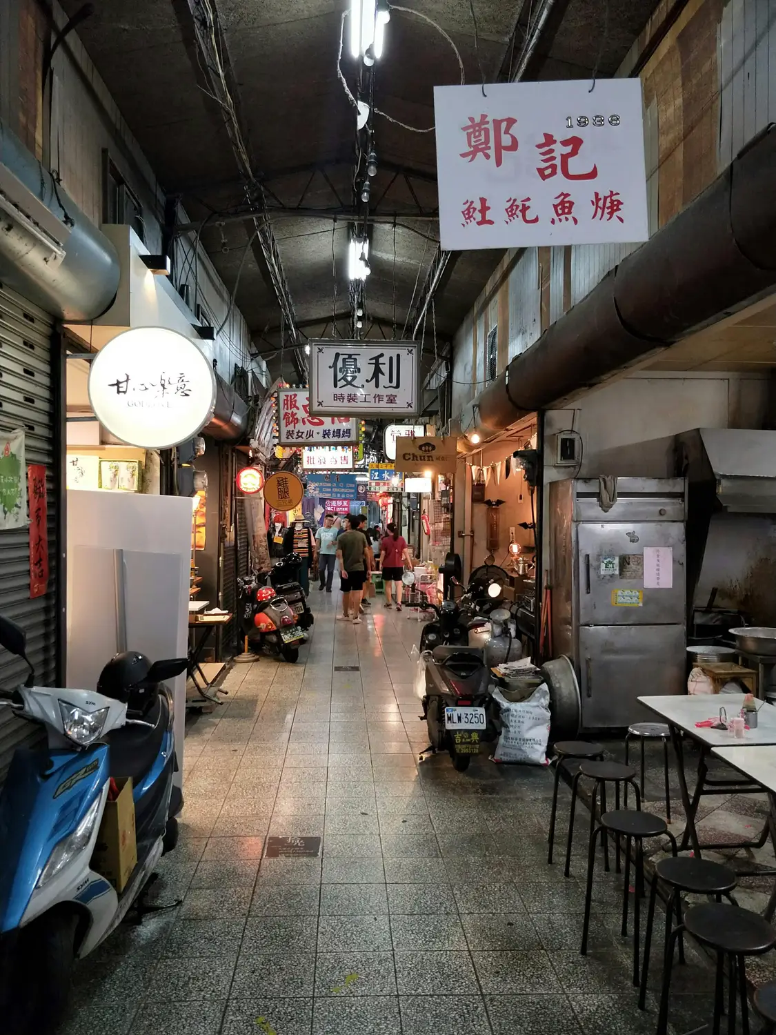 Dimly lit covered market alley in Tainan with hanging shop signs, parked scooters, and stools beside a traditional eatery dating to 1936