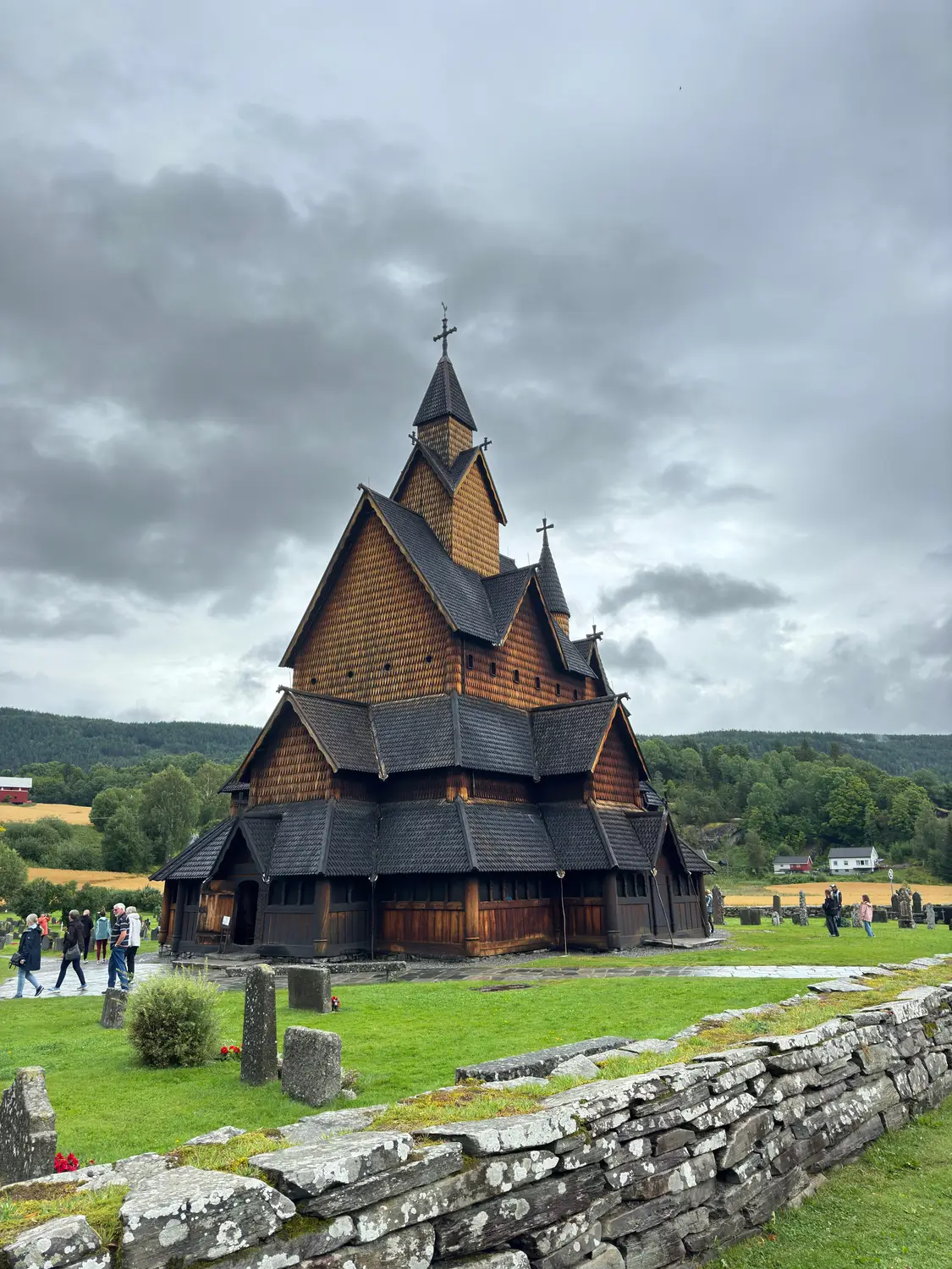 The dark timber tiers of Heddal Stave Church under an overcast sky, with visitors gathered on the green grounds.