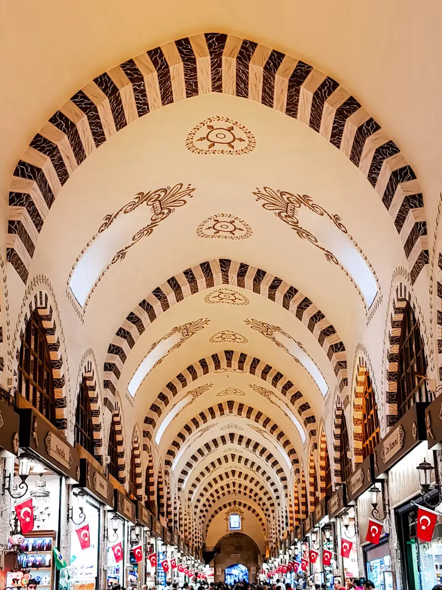 The vaulted, arched stone ceiling of a covered market corridor lined with shops and hanging Turkish flags.