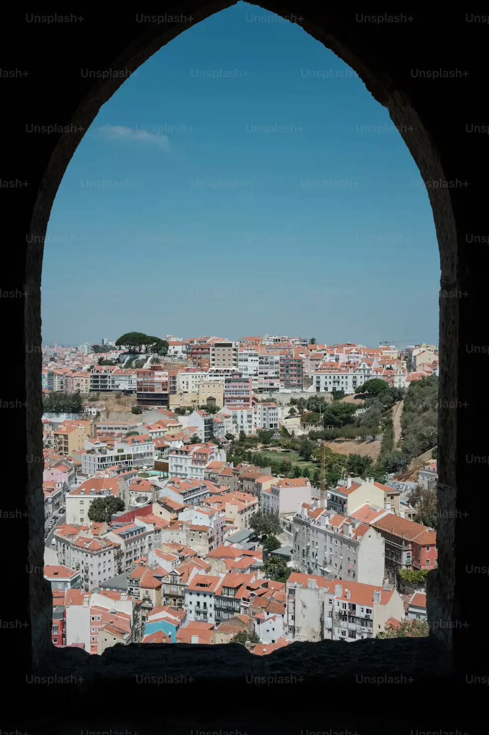 View of a city with terracotta rooftops framed by a tall stone archway against a clear blue sky.