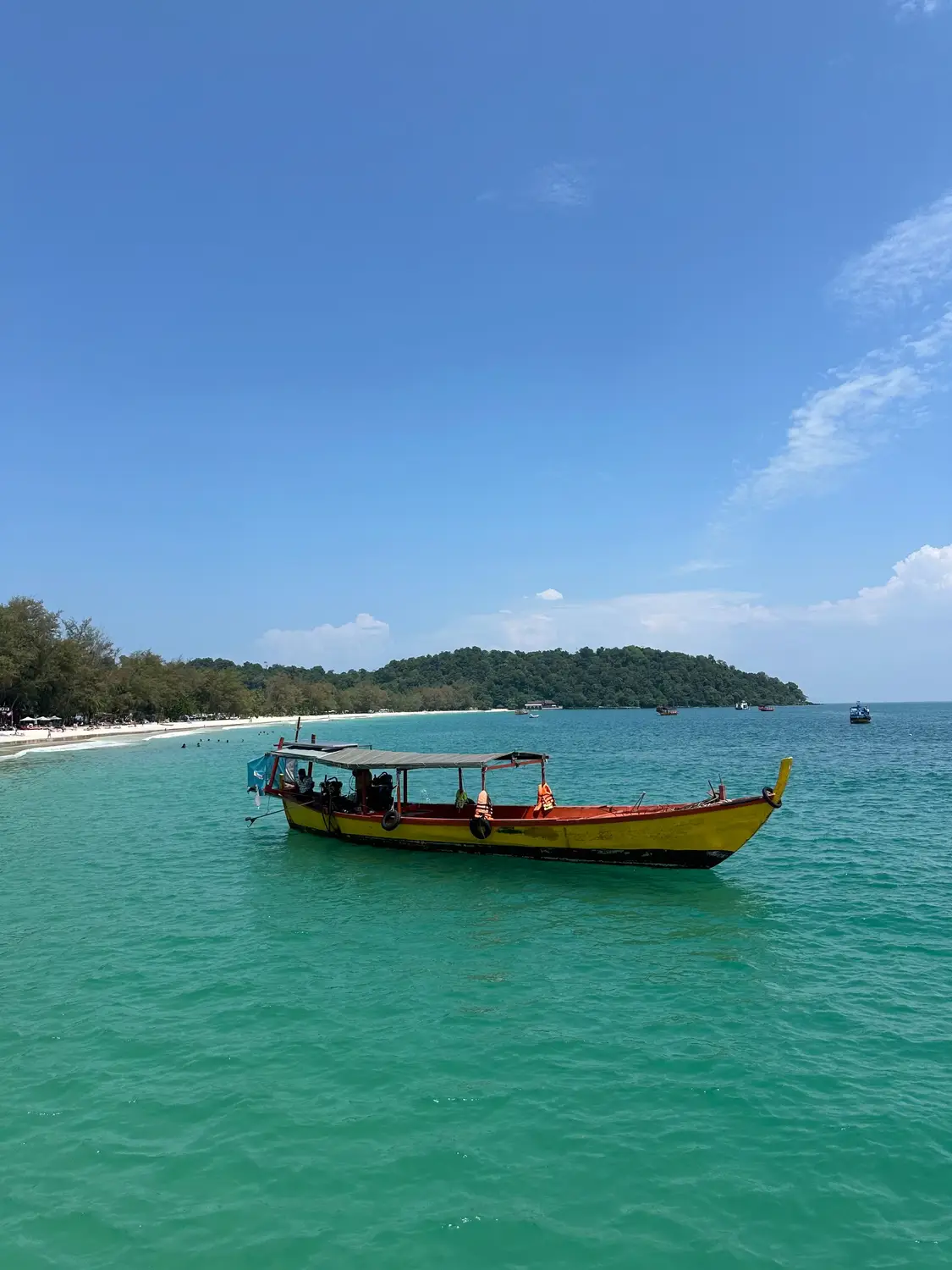 Yellow wooden longtail boat on turquoise sea water with white sand beach and lush green island in background, Koh Rong Cambodia