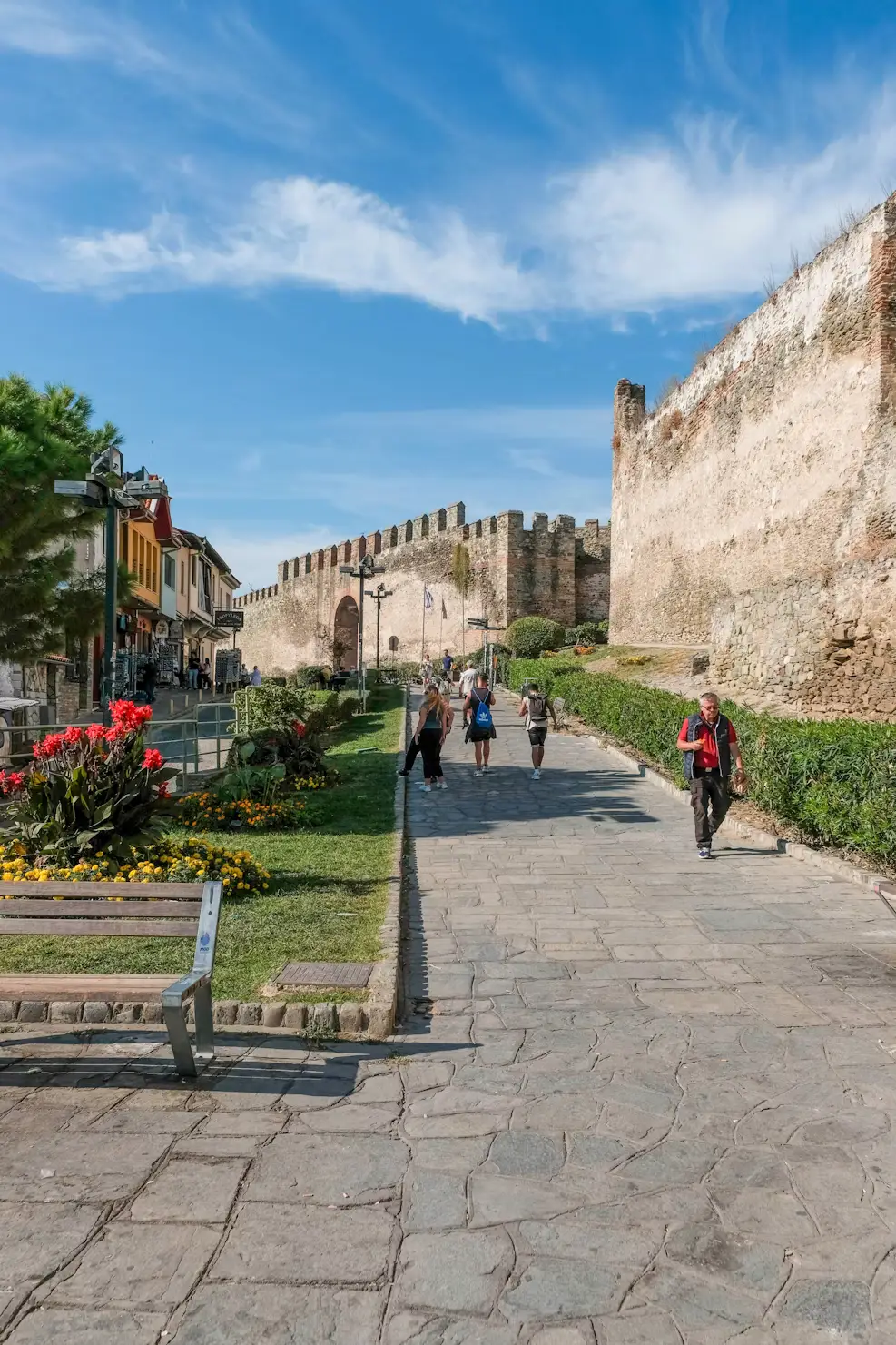 A stone pathway alongside an ancient fortress wall, with people walking and greenery lining the path.