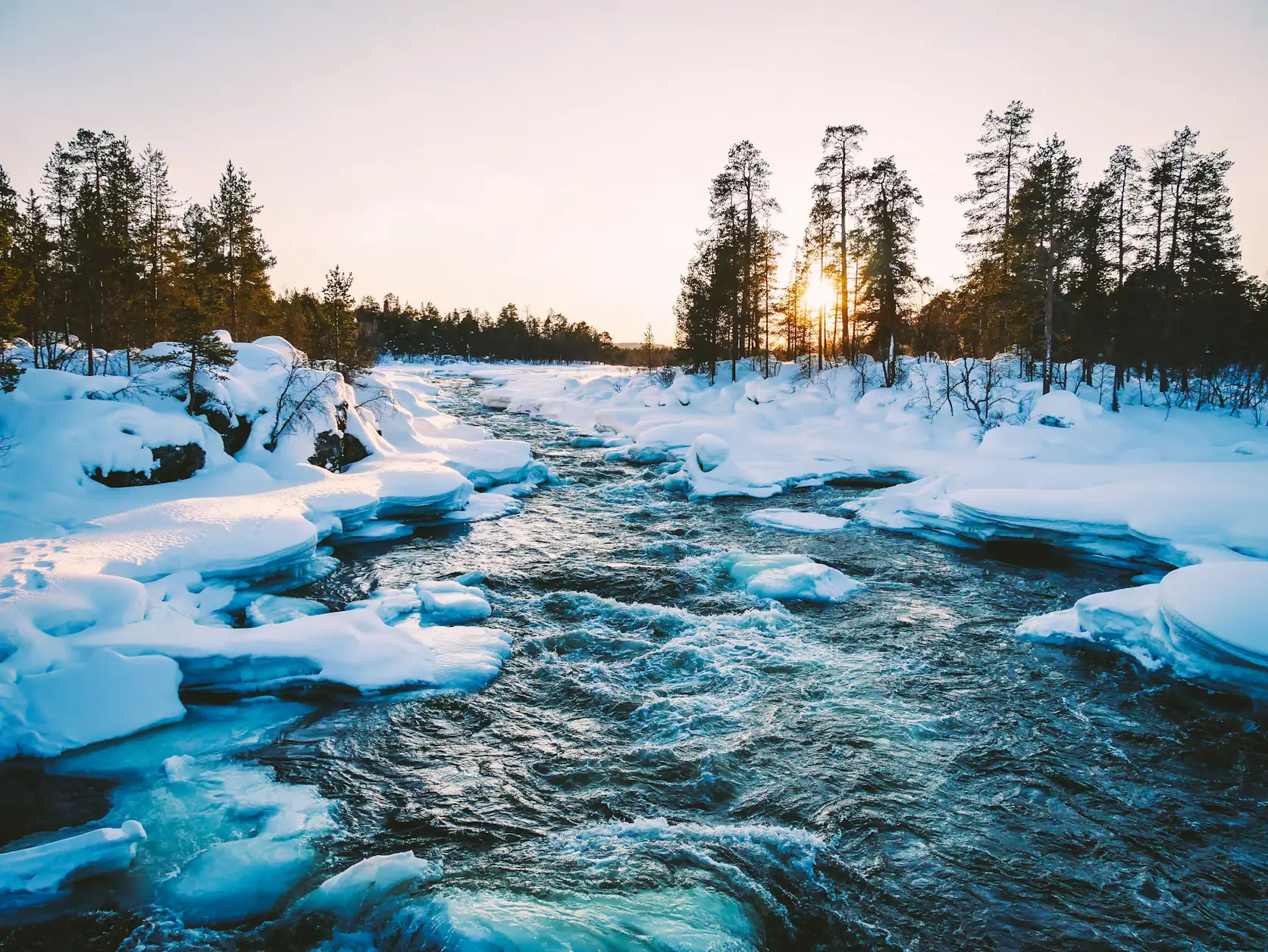 Snow-covered river flowing through a forest at sunset, with pine trees and icy banks.