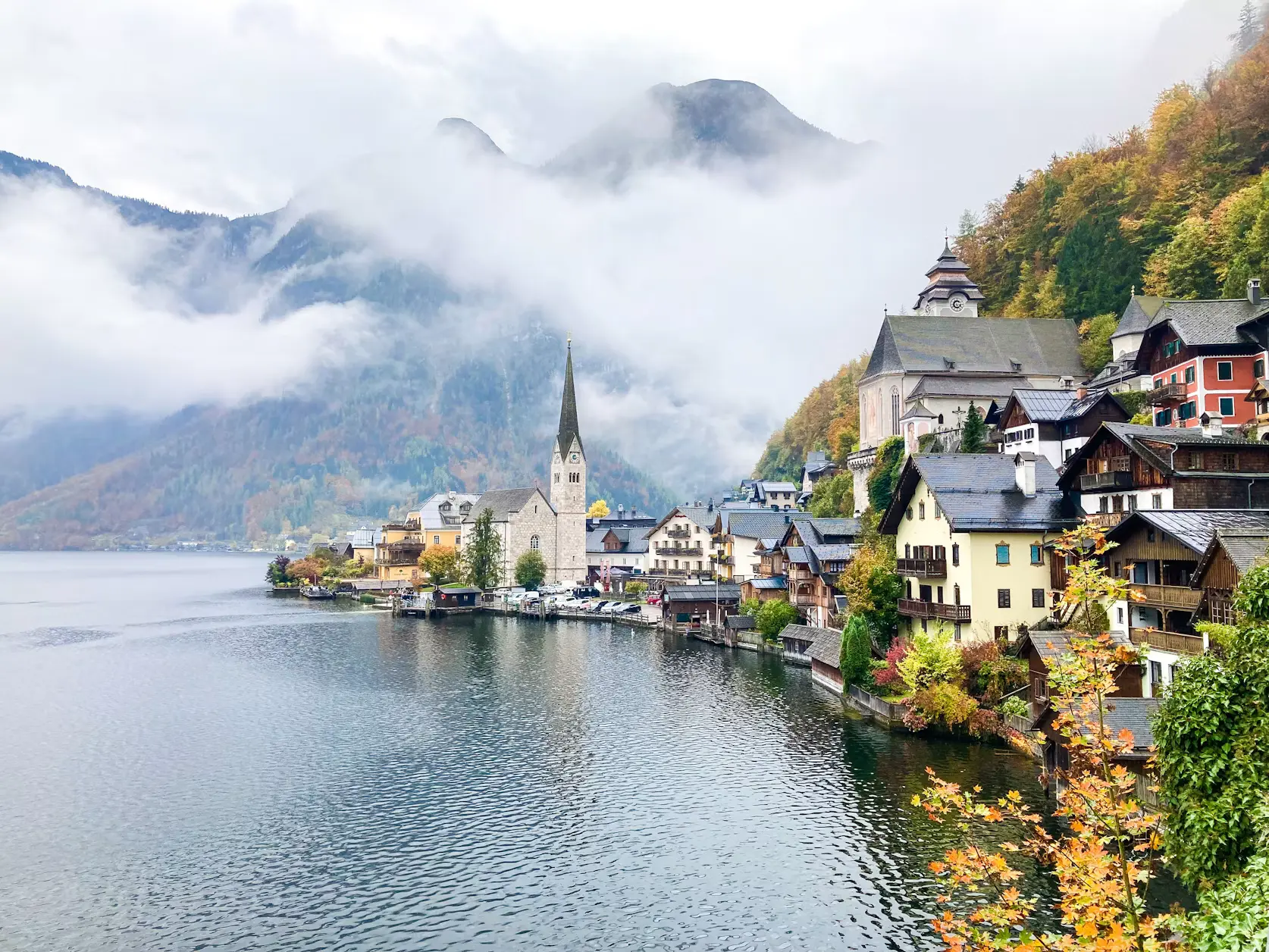 A misty lakeside village with colorful houses and a church steeple nestled against forested mountains, viewed across calm water with autumn-toned trees along the shore.