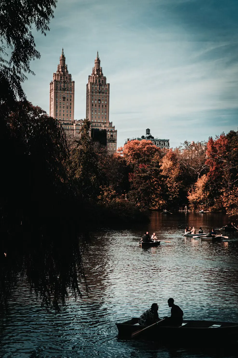 People rowing boats on the Central Park lake in autumn, with the San Remo's twin towers rising above the colourful trees.