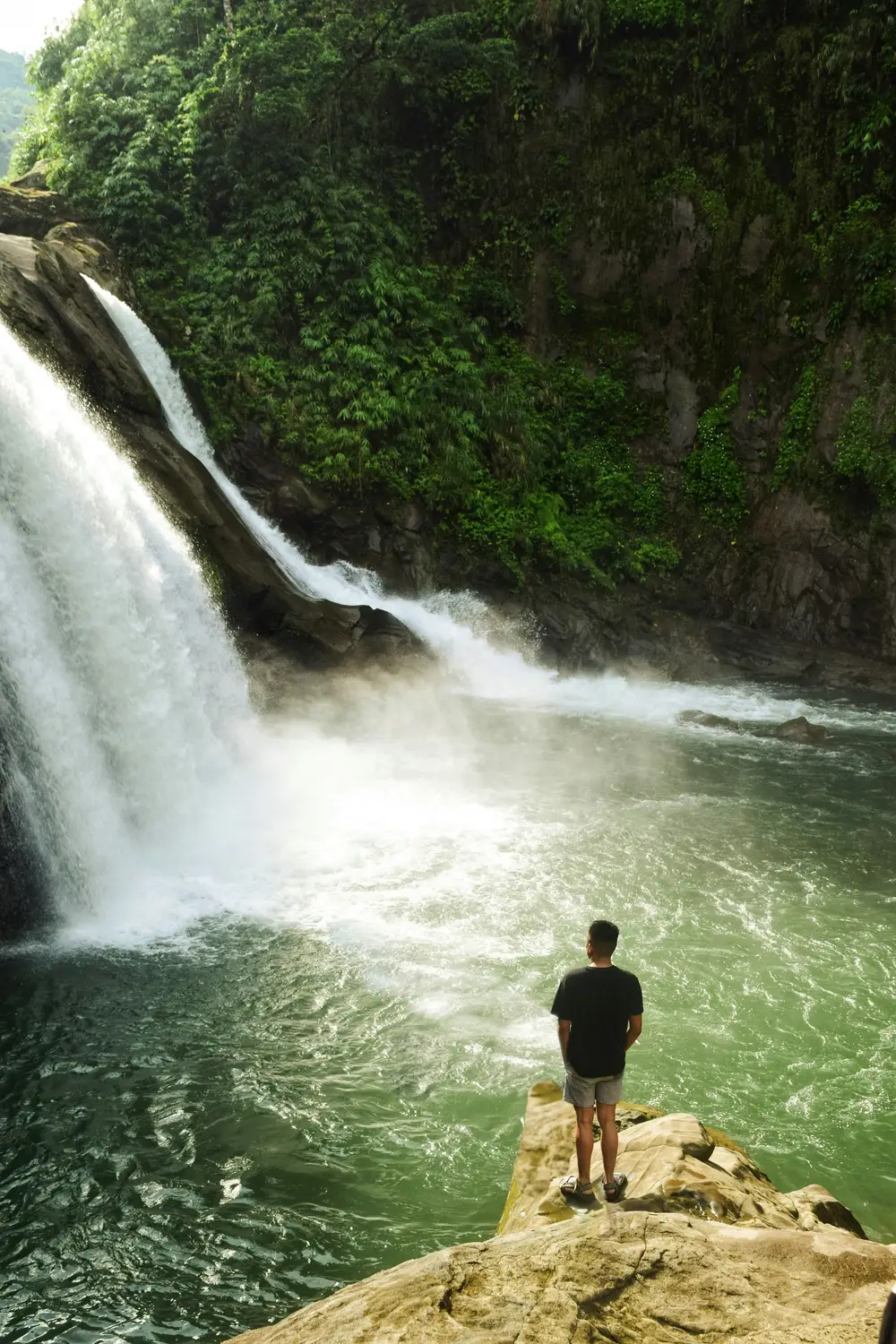 Person standing on a rock facing a waterfall surrounded by lush green forest in the Philippines.