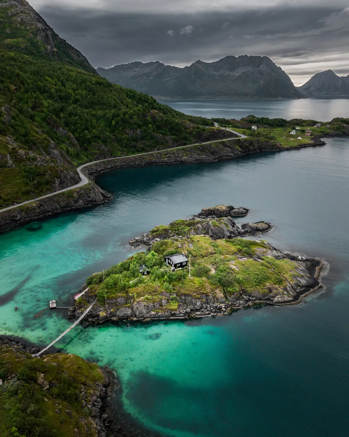 Aerial view of a coastal road curving past small green islands surrounded by shallow turquoise water under a moody sky.