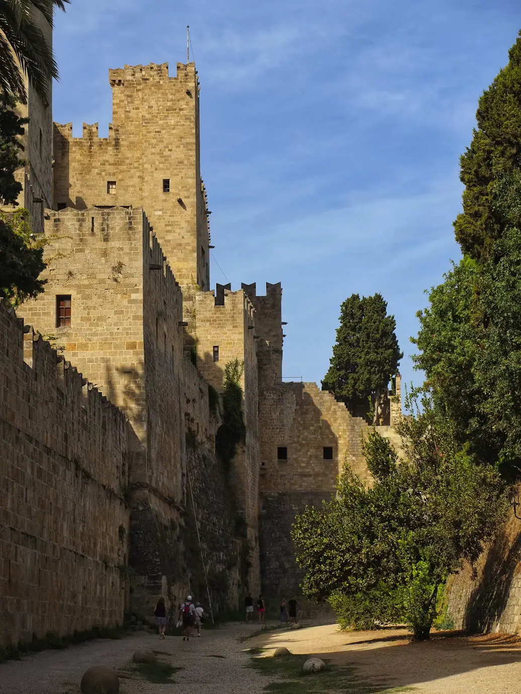 A medieval stone fortress wall and tower, with trees and a footpath running alongside it under a blue sky.