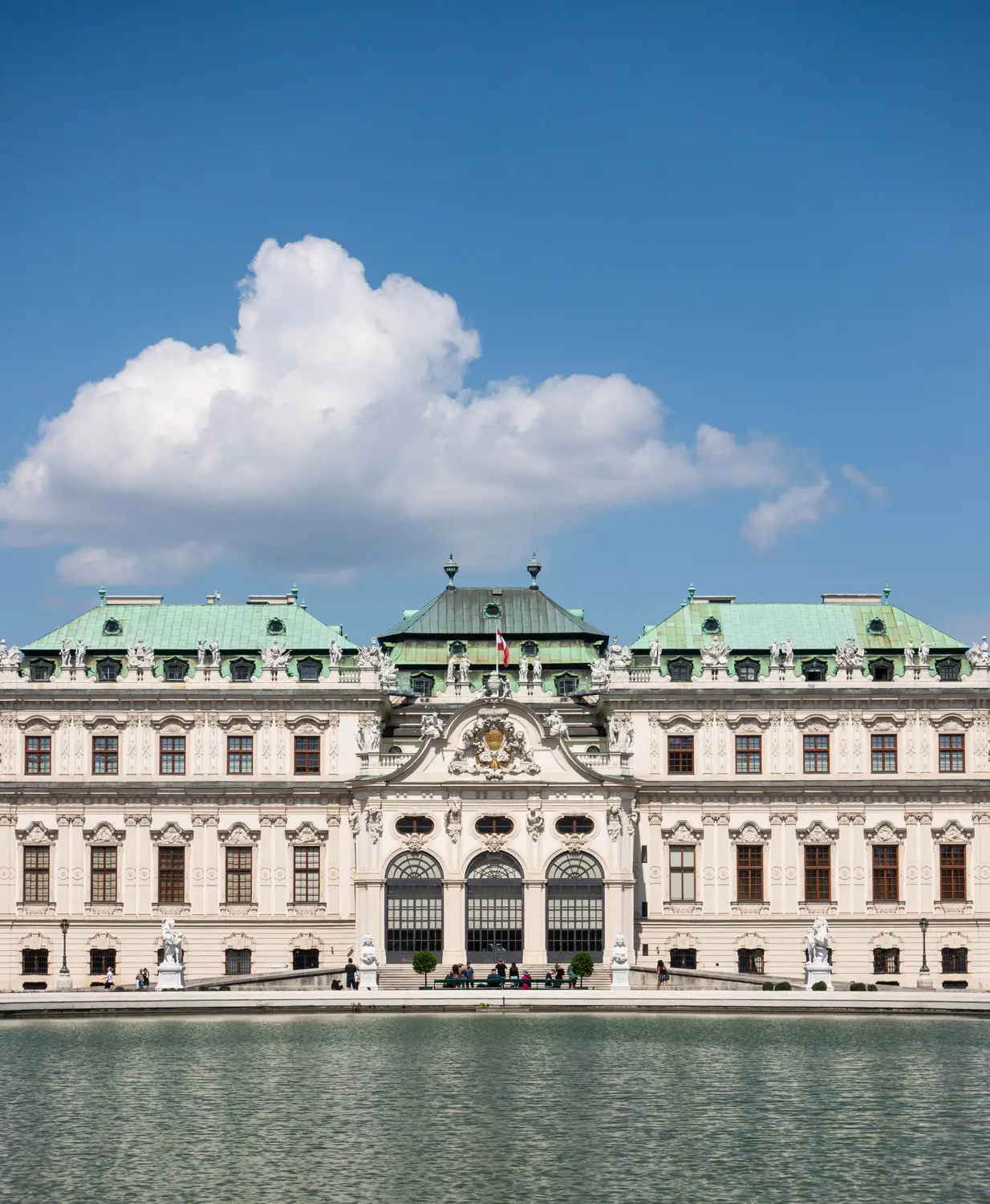 A grand baroque palace with ornate facades and a green dome, reflected in a still pool under a blue sky with white clouds.