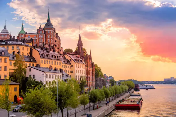 Colorful old-town buildings and a church spire in Stockholm's Södermalm district at sunset, overlooking the waterfront with boats along the promenade.