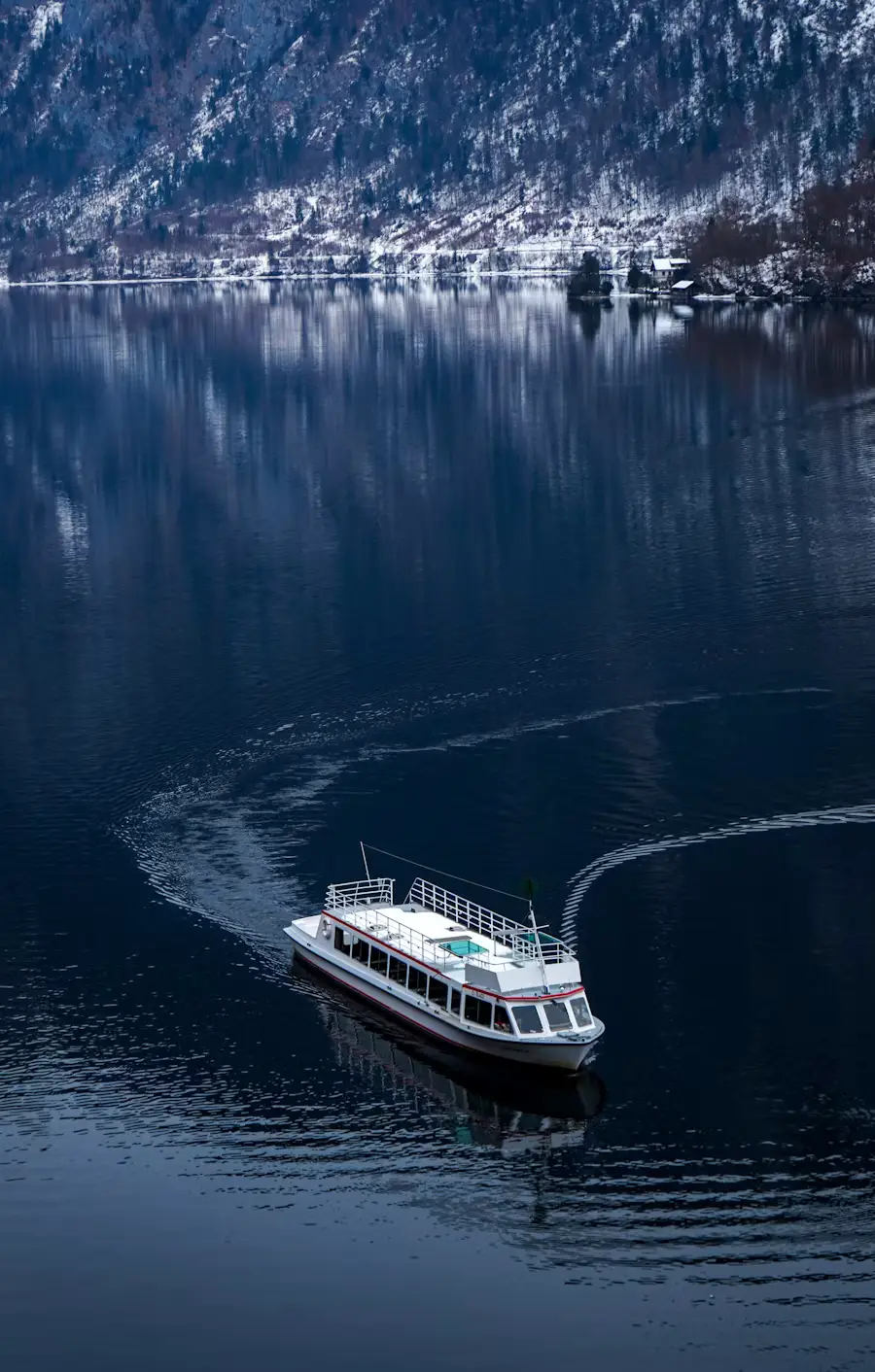 A small tour boat leaves a wake as it crosses a still lake at dusk, framed by snow-dusted mountains reflected in the dark water.