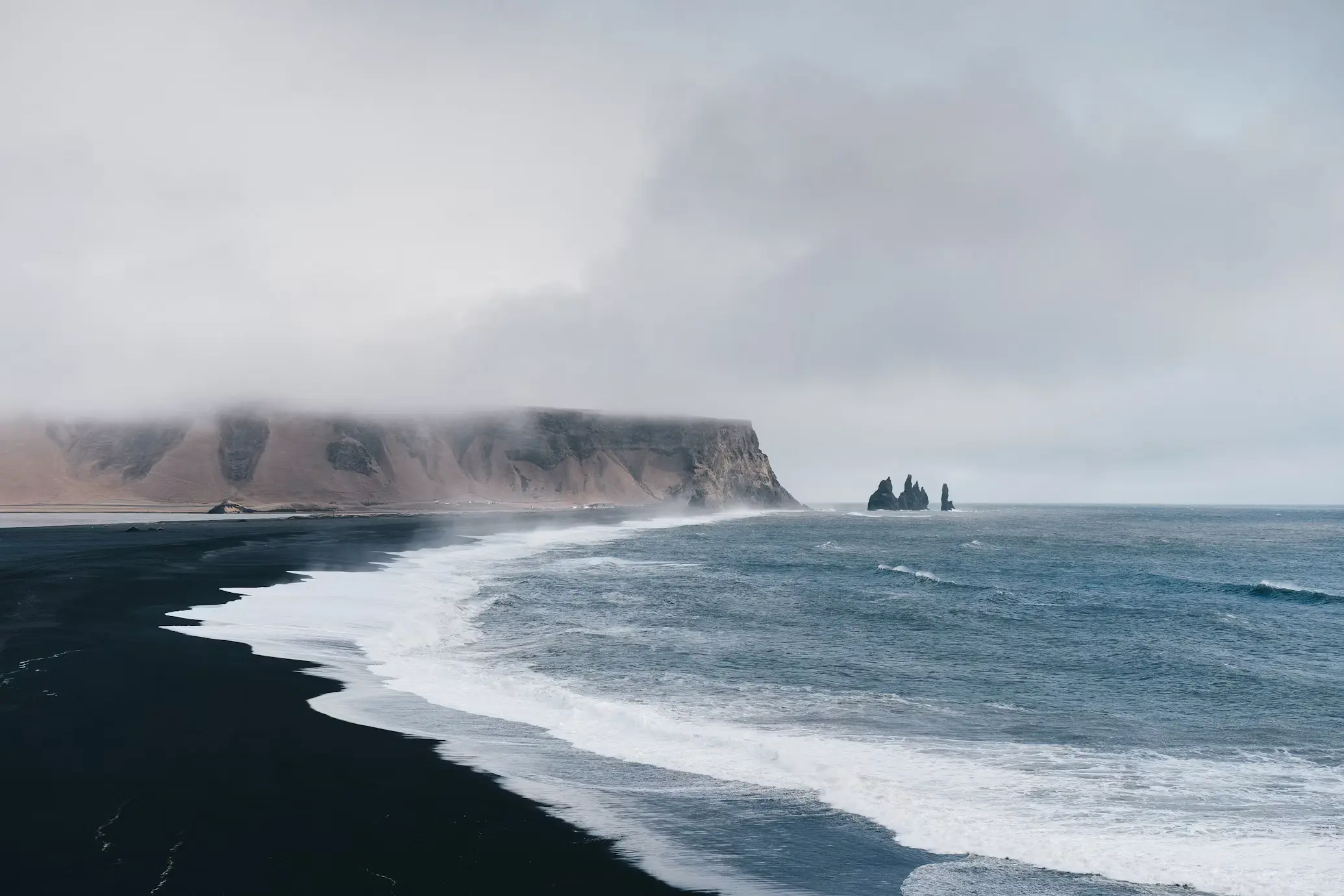 Black sand beach with crashing waves along a misty coastline, sea stacks visible in the distance, Iceland.
