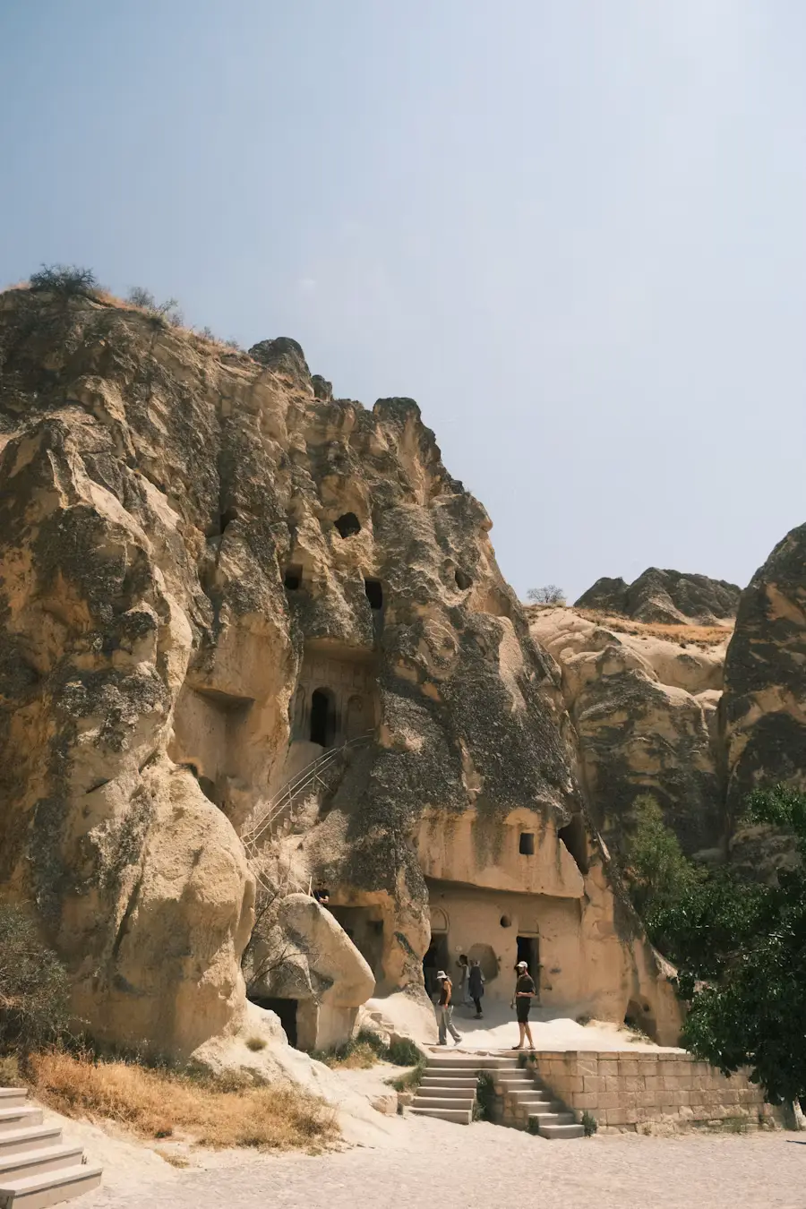Ancient cave dwellings carved into a rocky cliff face, with a staircase leading up to the openings, in Cappadocia, Turkey.