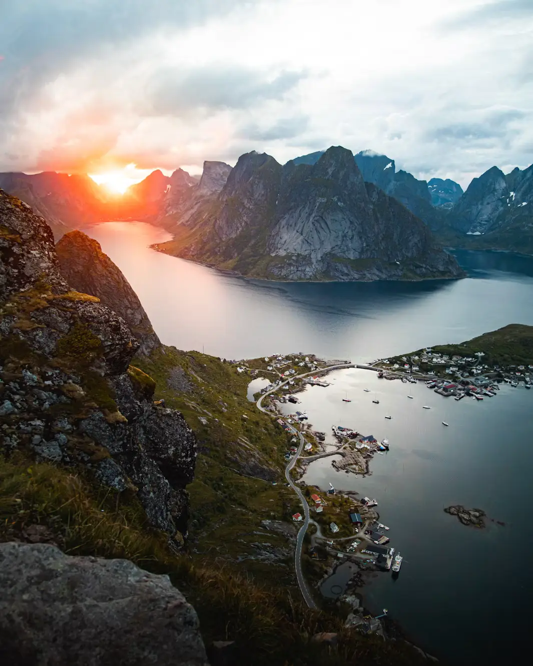 View from a mountainside over the village of Reine in Lofoten, with the sun setting behind jagged peaks surrounding the fjord.