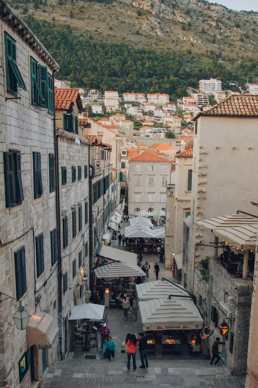 Aerial view of a narrow old-town street lined with shops, awnings, and pedestrians, hillside buildings in the background.