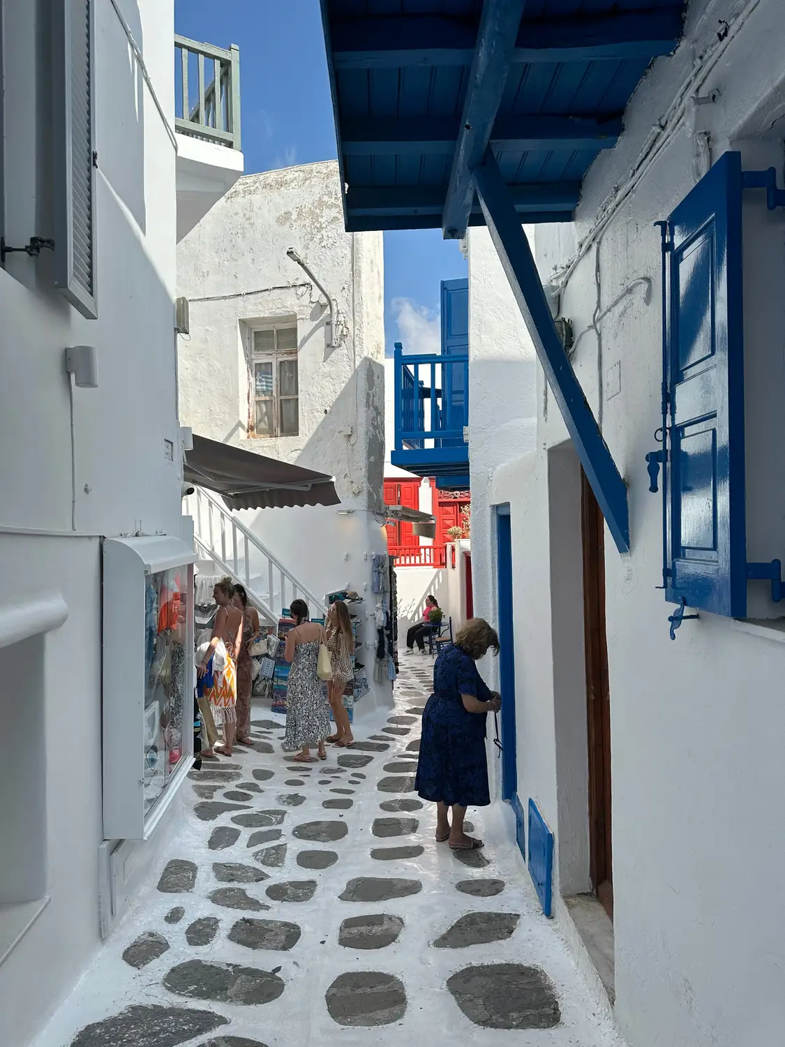 A narrow whitewashed alley in Mykonos with blue-shuttered windows and people walking past shops.