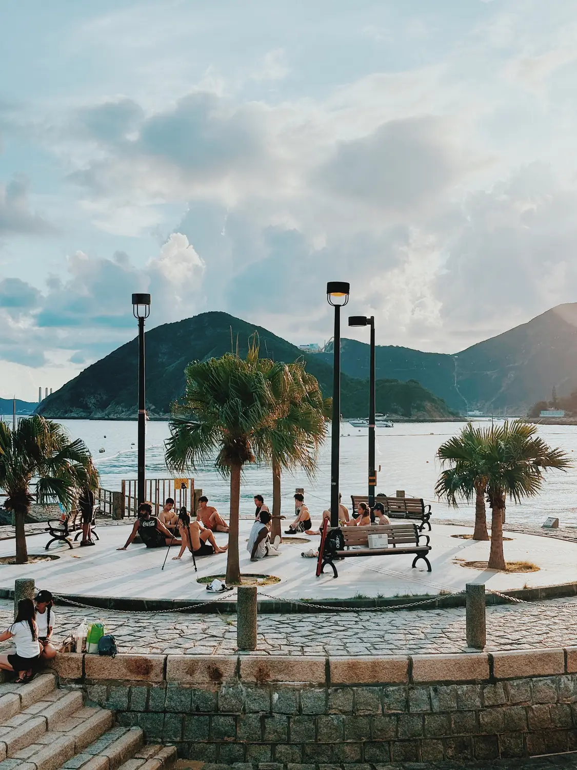 Palm-lined waterfront promenade with people relaxing and green hills across the water in Hong Kong.