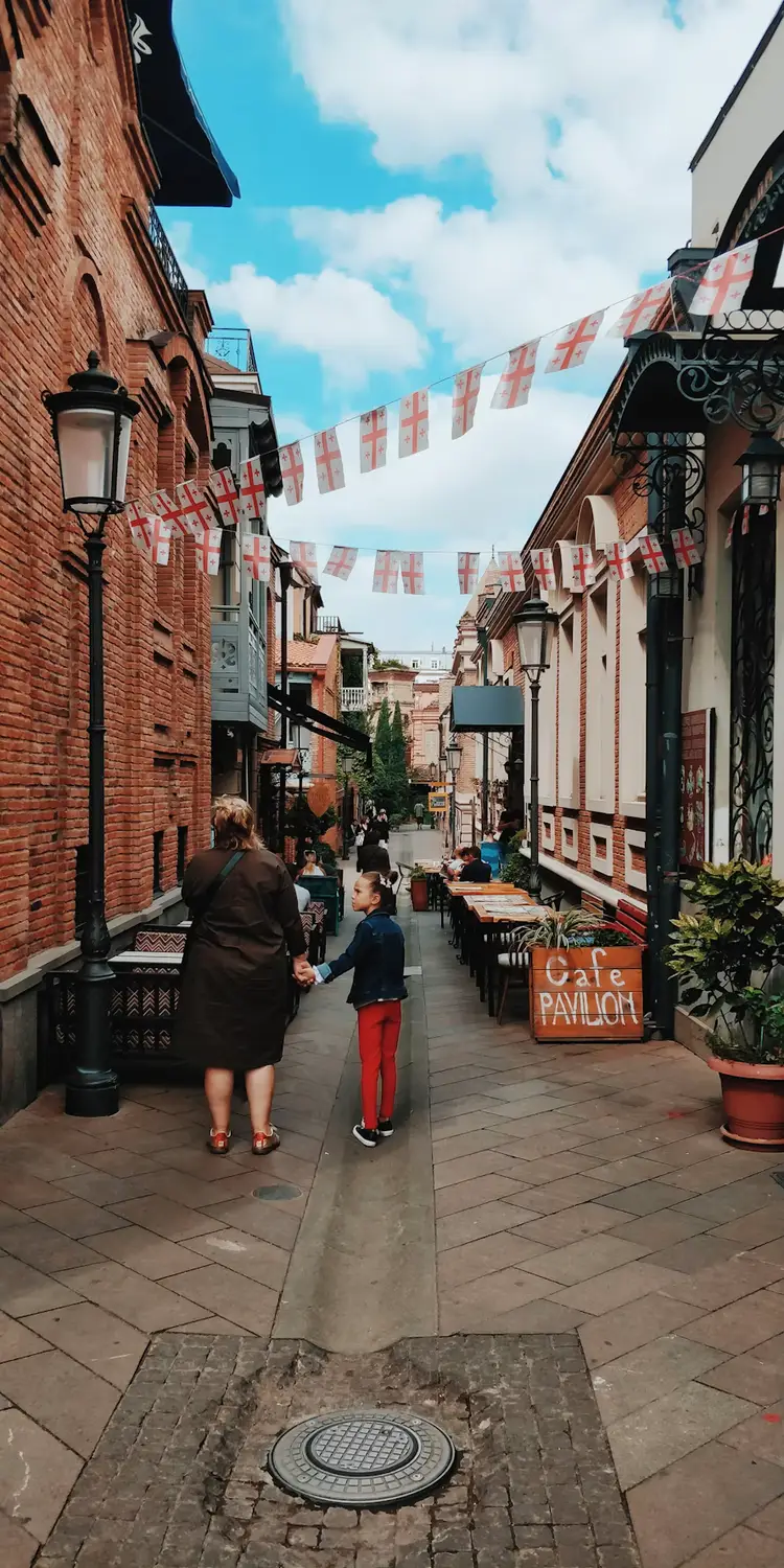 A narrow cobblestone street lined with historic brick buildings, cafés, and Georgian flags, with a woman and child walking beneath a bright blue sky.