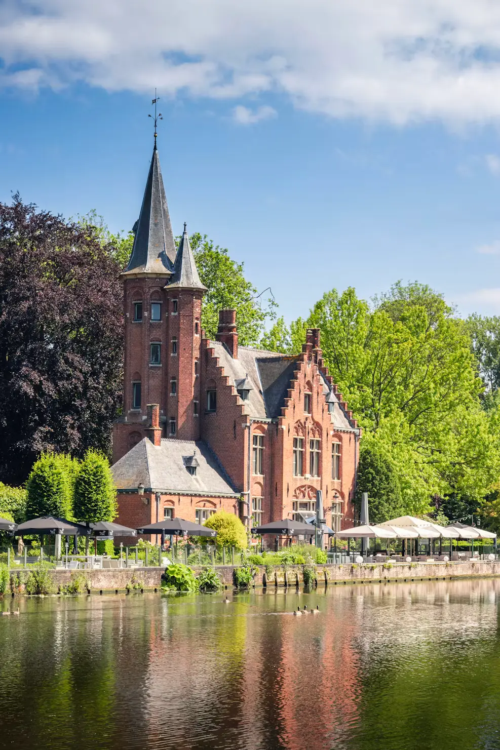 The red-brick Lock House on Bruges' canal, its stepped gables and turret reflected in calm water beside outdoor café umbrellas.