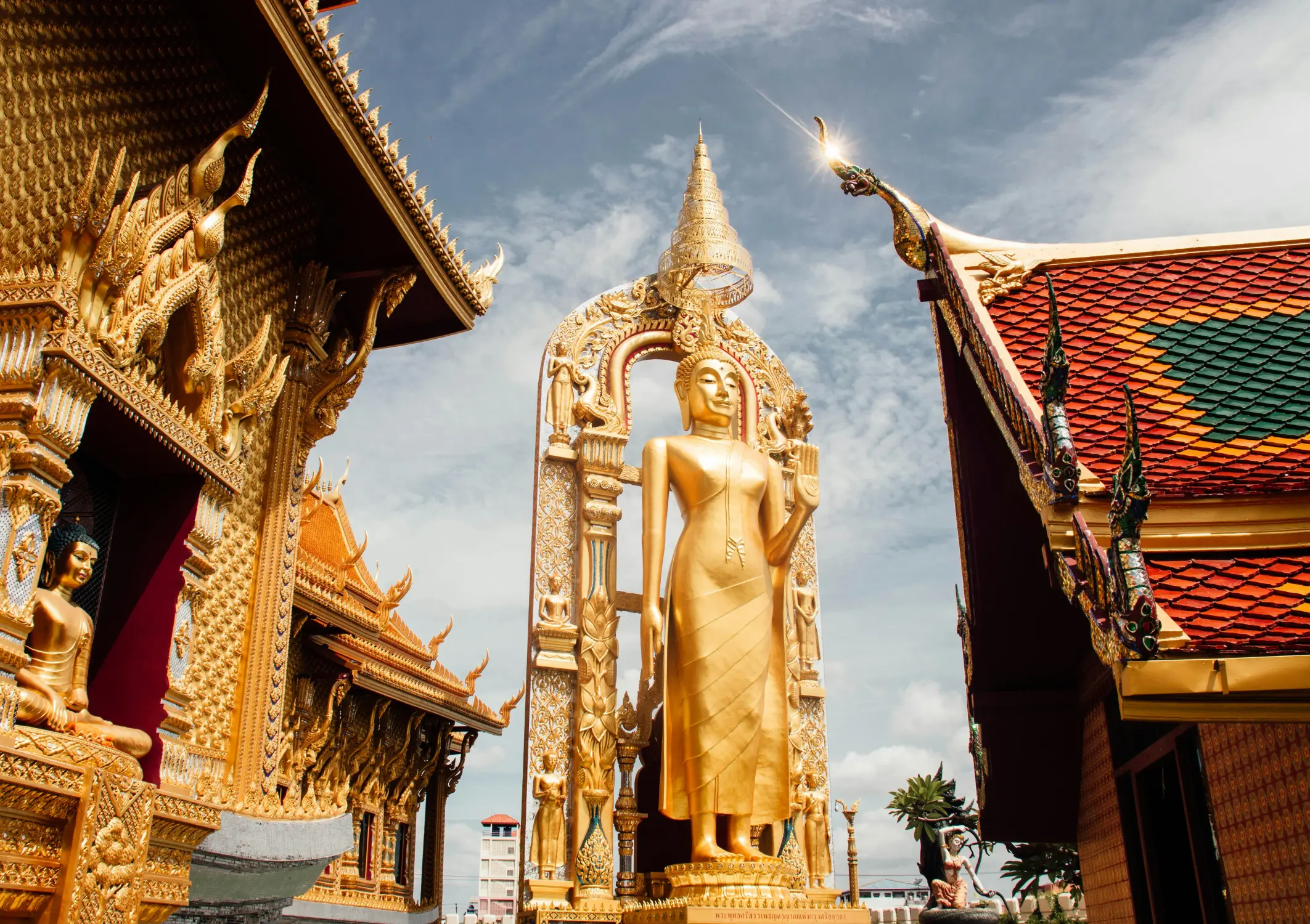 Golden standing Buddha statue displayed between ornate Thai temple buildings, with intricate gold detailing, colorful tiled roofs, and a bright blue sky.