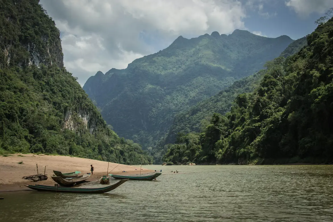 Wooden boats moored along a river beneath lush green forested mountains in Laos.