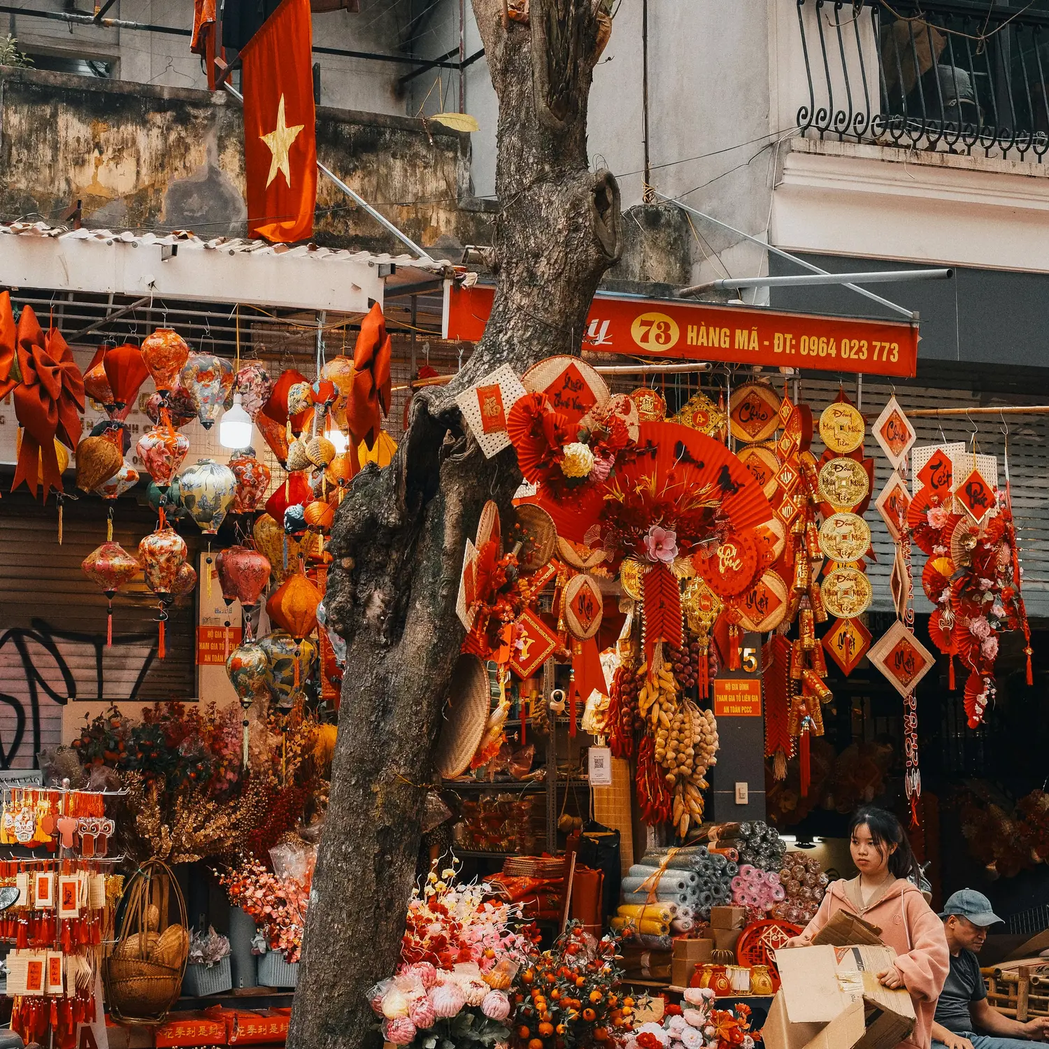 Festive red Tet decorations, lanterns, and traditional ornaments displayed at a shop on Hang Ma Street in Hanoi's Old Quarter, Vietnam, with a Vietnamese flag and vendors below.