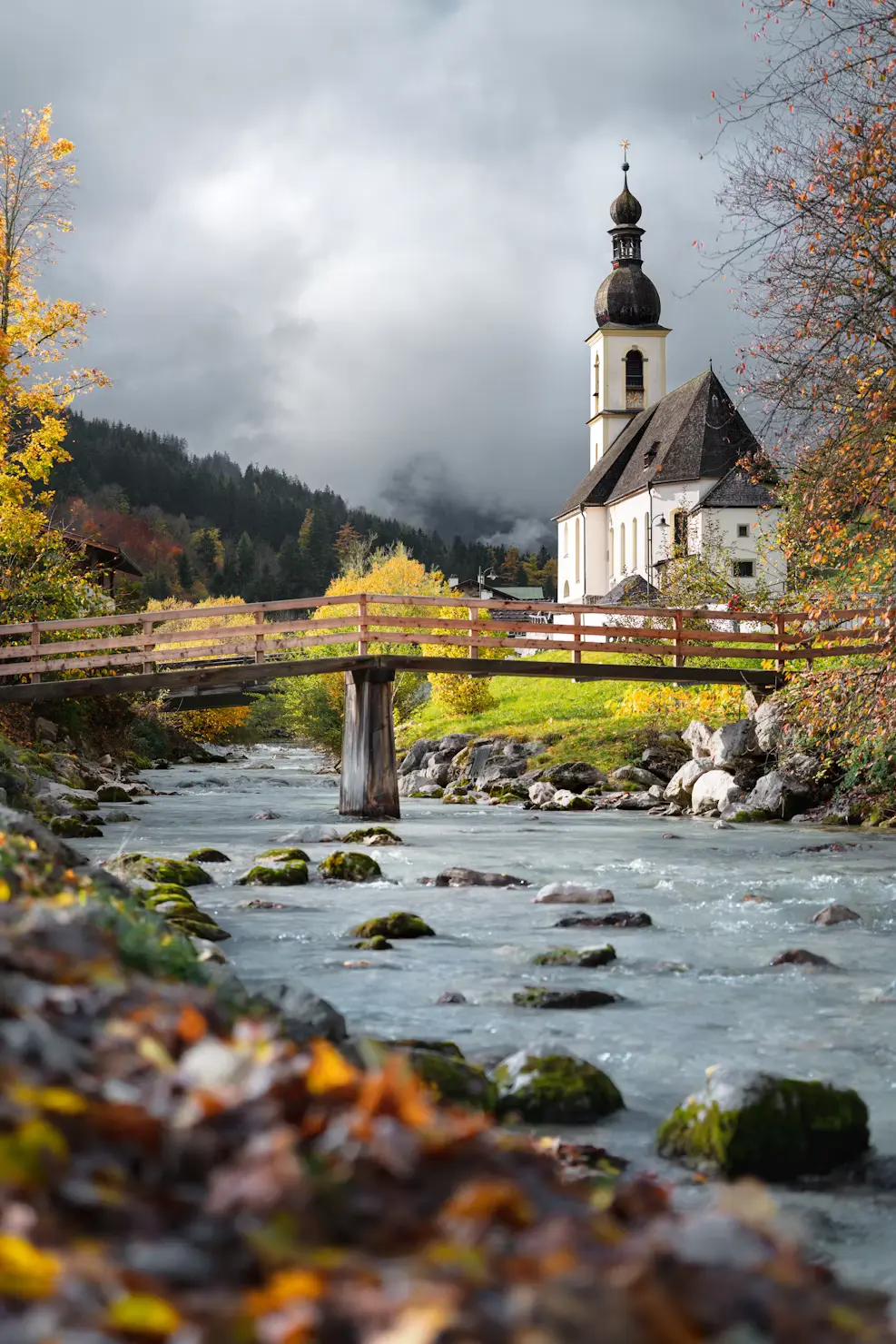 White pilgrimage church beside a mountain stream with a footbridge and autumn foliage