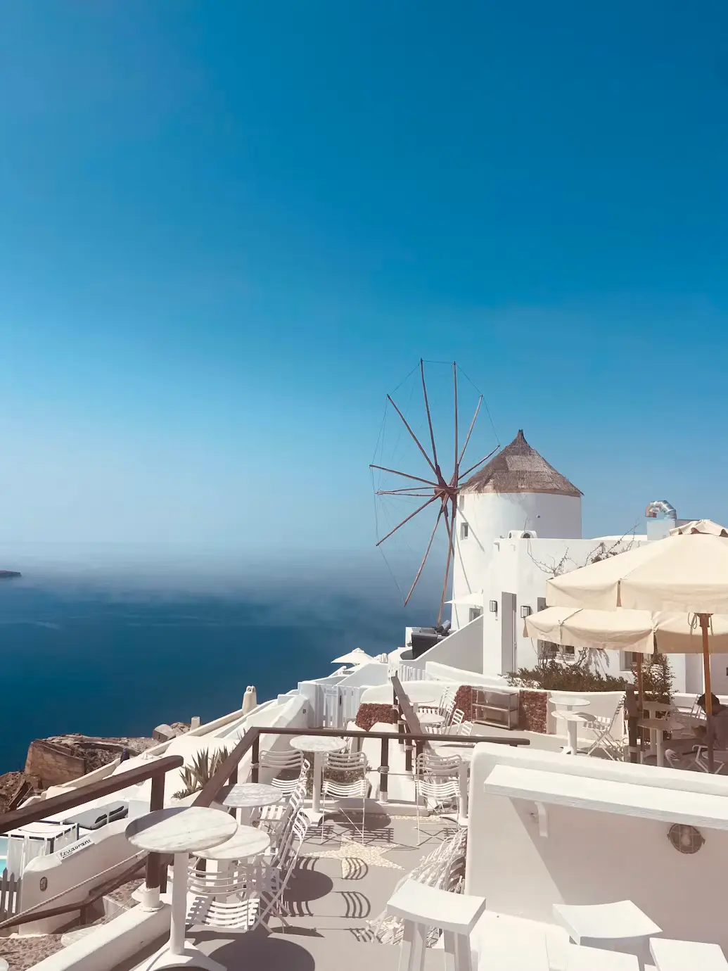 A traditional white windmill and outdoor café seating overlooking the sea in Santorini, Greece, under a clear blue sky.