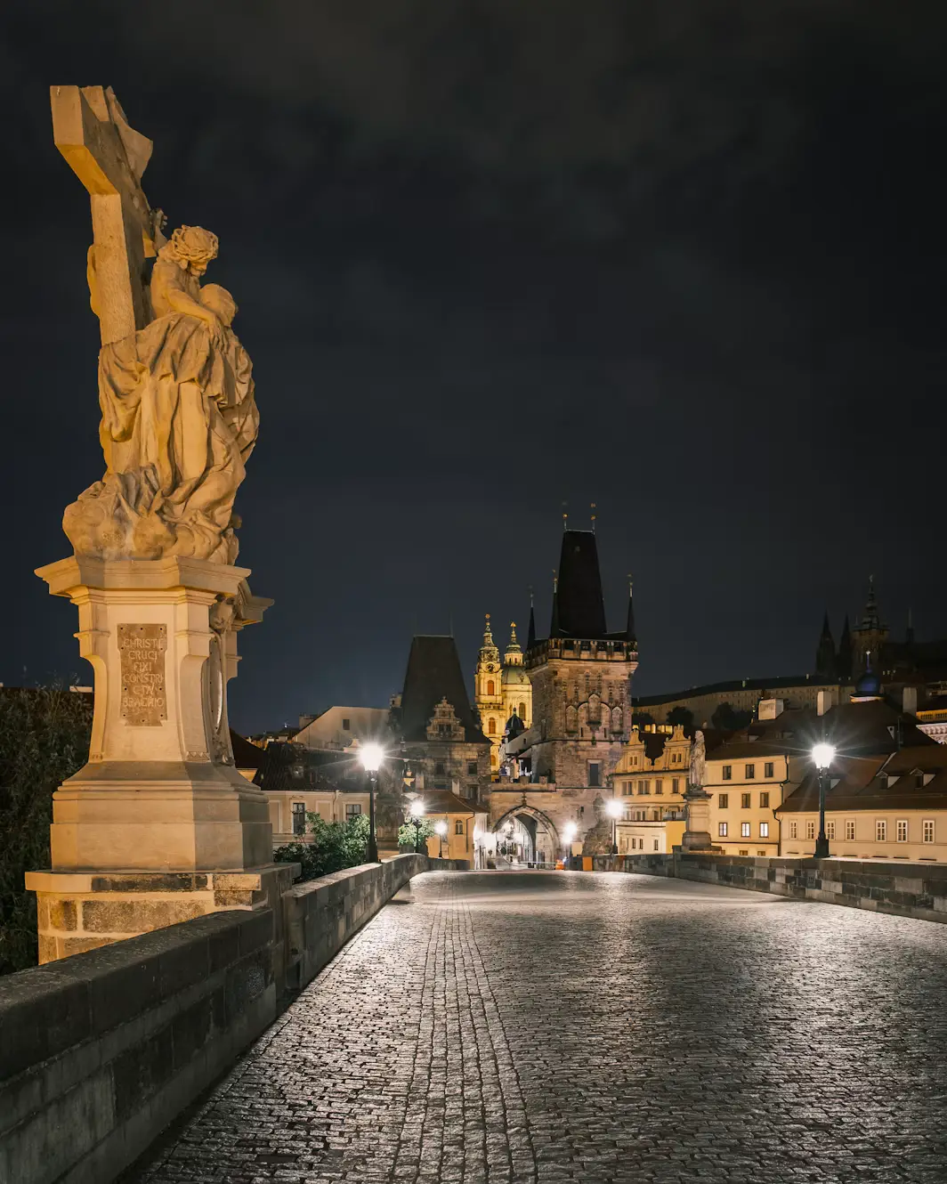 Charles Bridge at night, lined with statues and lamplight, with Old Town Bridge Tower in the distance