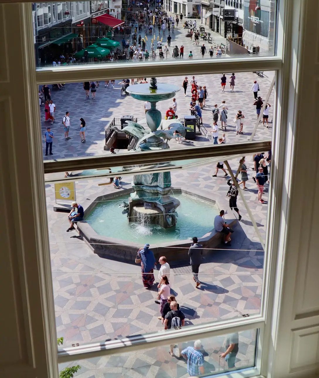 View through a window overlooking a fountain and plaza filled with people in Copenhagen