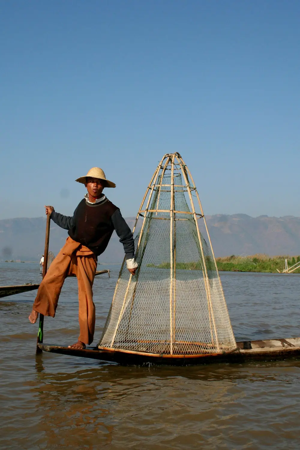 Traditional Intha leg-rowing fisherman standing on wooden boat with conical bamboo fish trap on Inle Lake, Myanmar