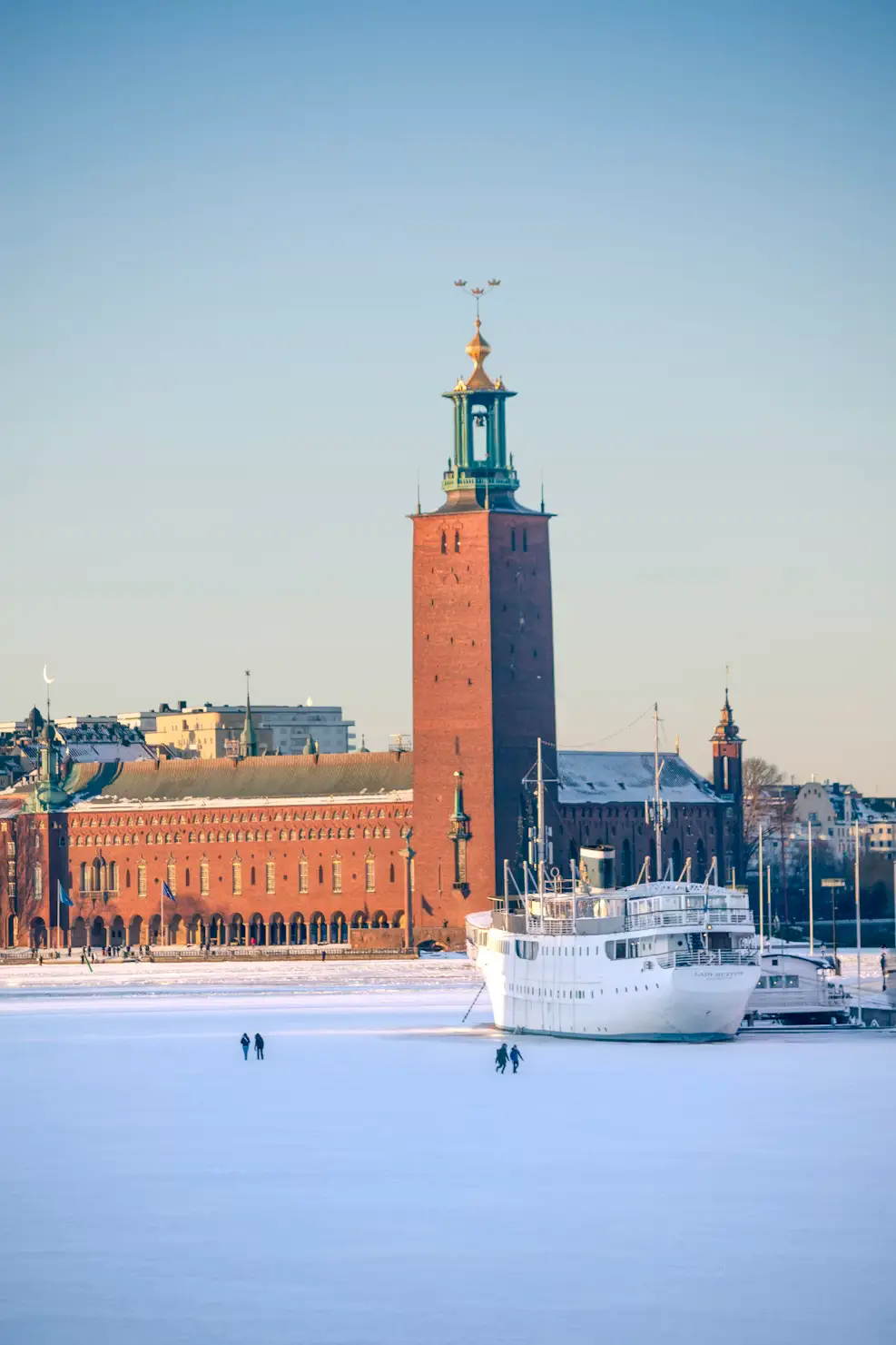 Stockholm City Hall's tower rising above a frozen, snow-covered waterway, with a boat docked nearby.