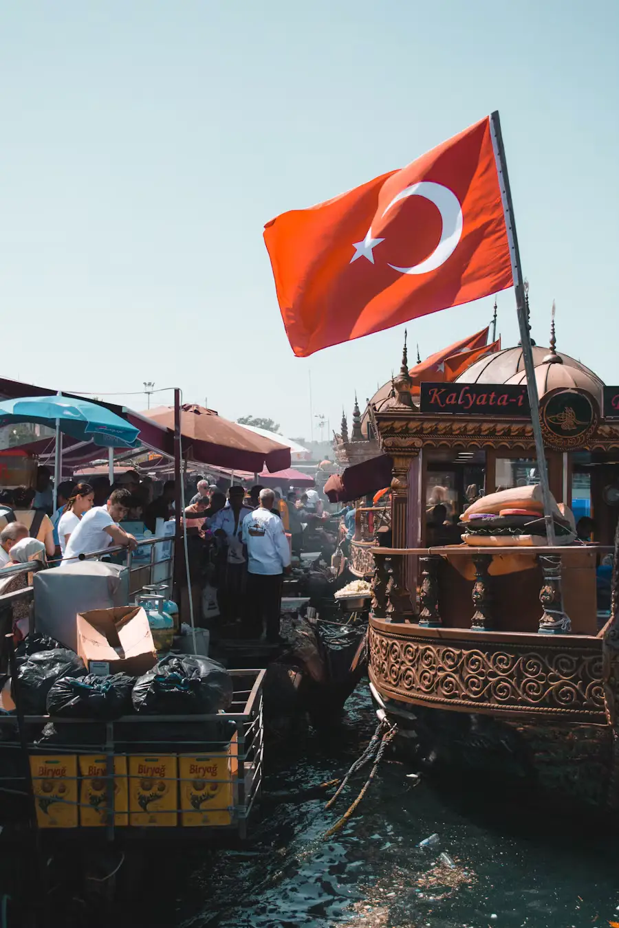 A Turkish flag waving above a bustling waterside market with boats and vendor stalls.