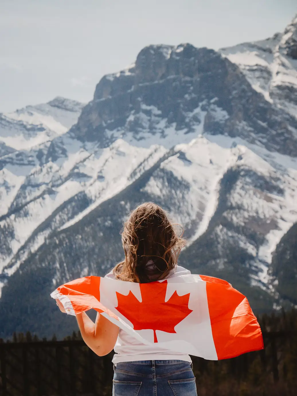 A woman seen from behind holding a Canadian flag around her shoulders, facing snow-capped mountains.