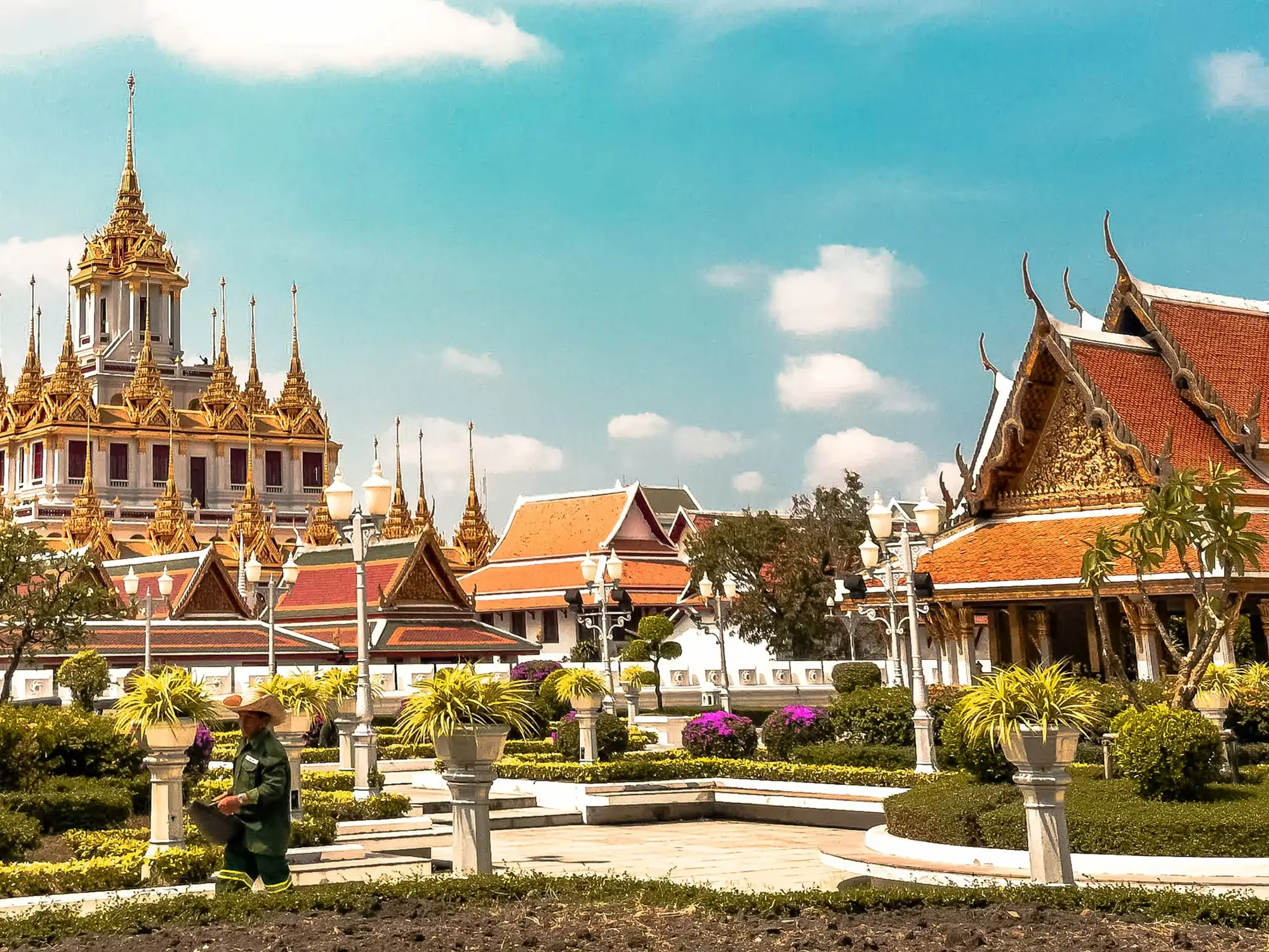 Ornate Thai Buddhist temple complex with traditional red-and-gold roofs, a tall golden central pavilion, manicured gardens, and a bright blue sky with scattered clouds.