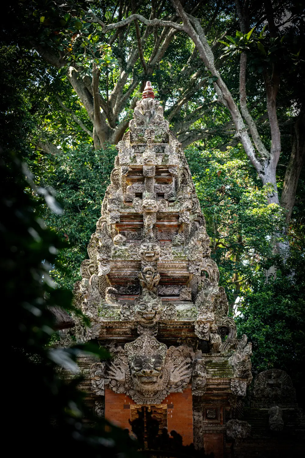 Ancient tiered Balinese stone temple tower with intricate carvings, surrounded by dense tropical forest and dappled sunlight in Bali, Indonesia.