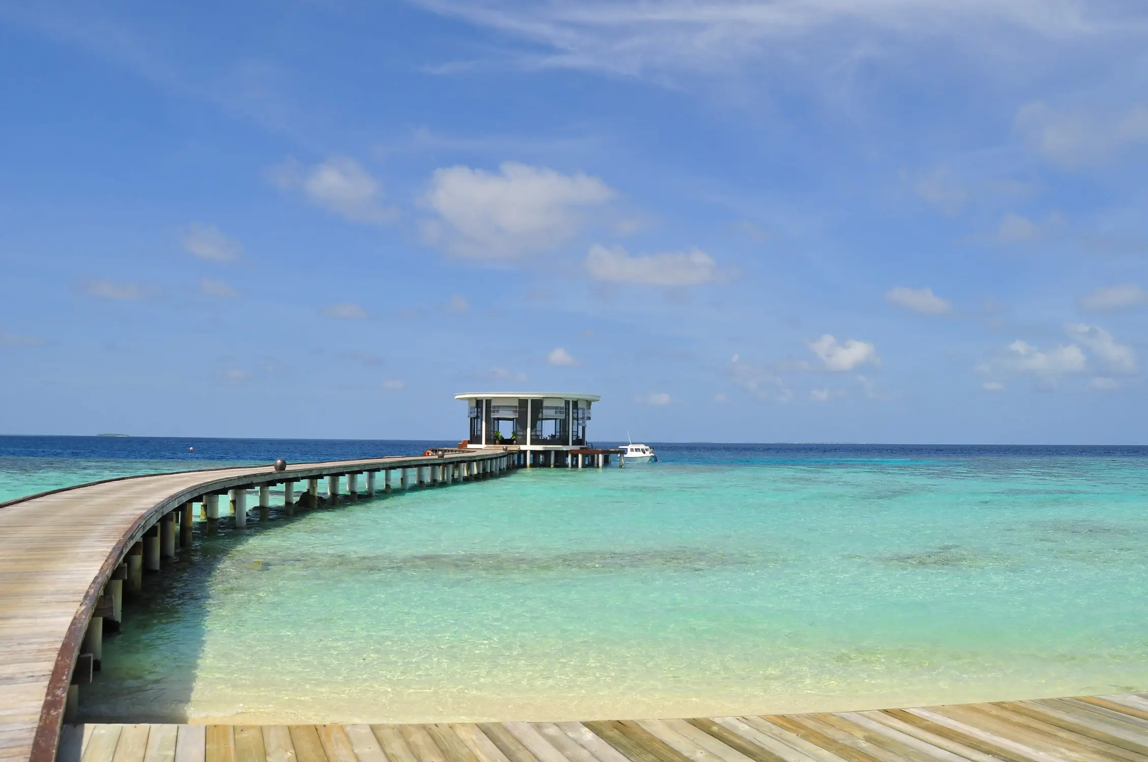Curved wooden jetty boardwalk extending over clear turquoise lagoon to overwater pavilion restaurant at Maldives luxury resort
