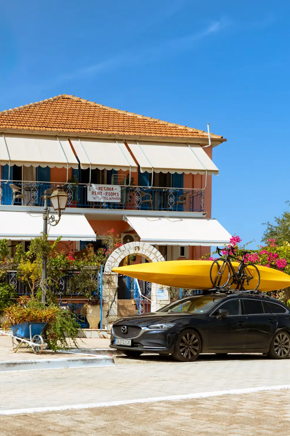 A traditional Greek building with balconies and hanging plants, with a car carrying a kayak parked on the street below.